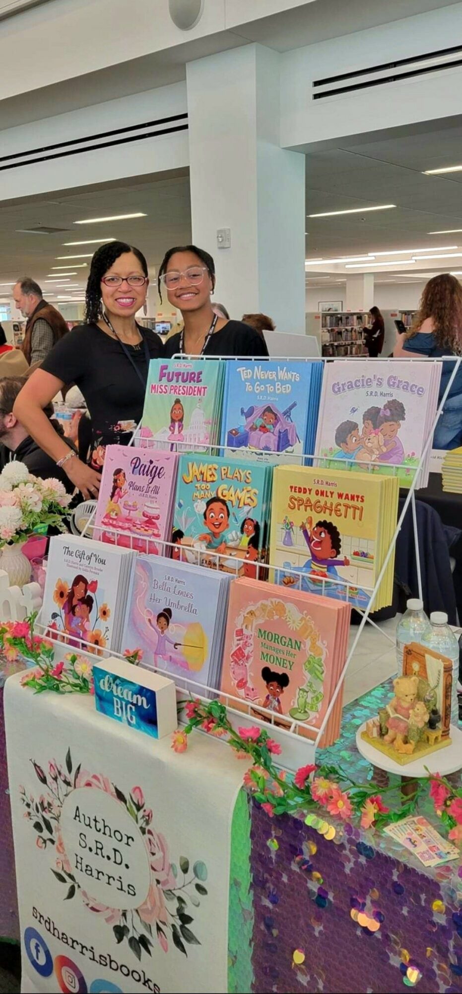 Two women standing behind a table with children's books and a cake, in a bright indoor space, smiling.