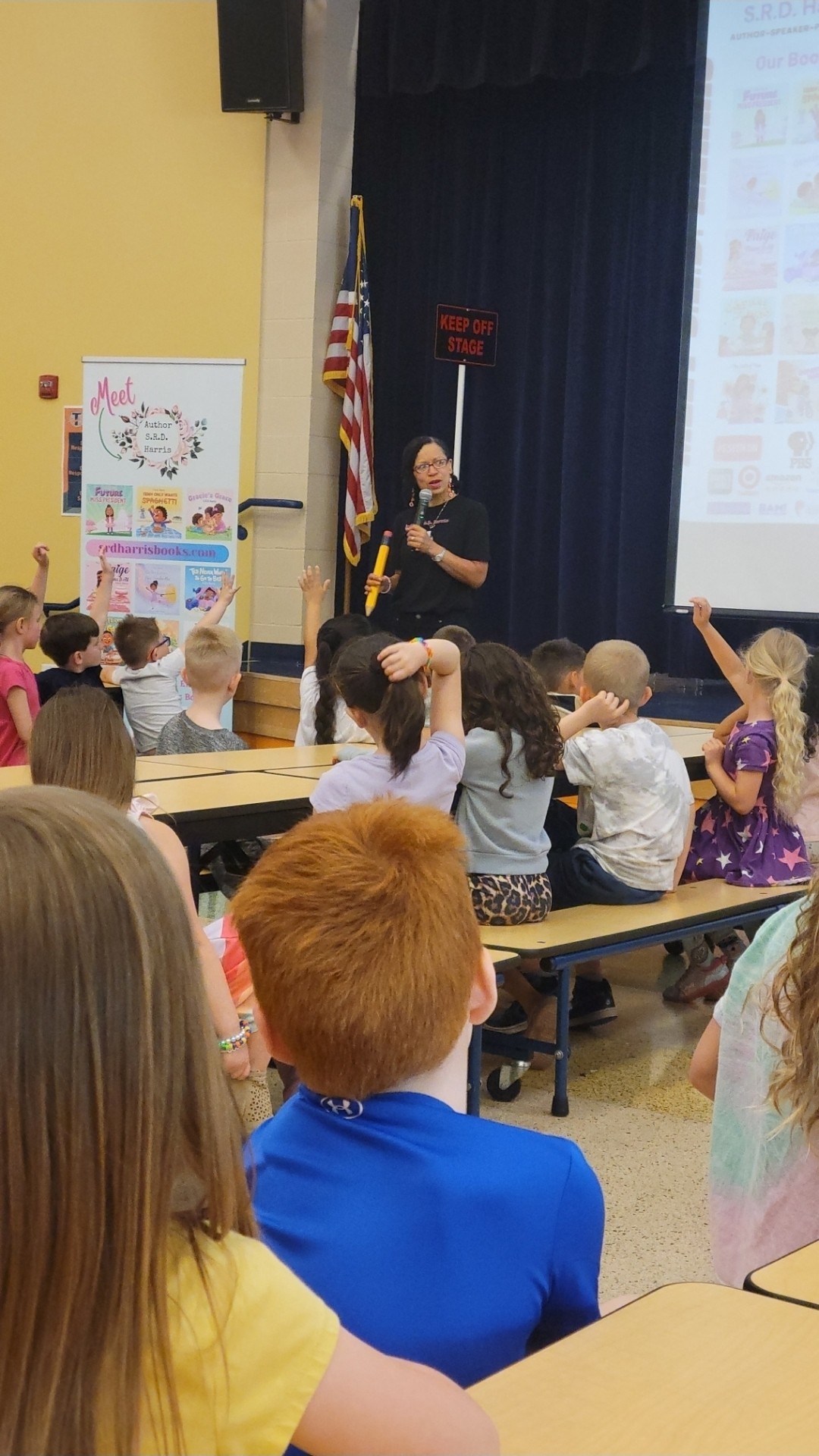 Person standing on stage holding a fire axe in front of seated children, with a blue curtain and American flag behind.