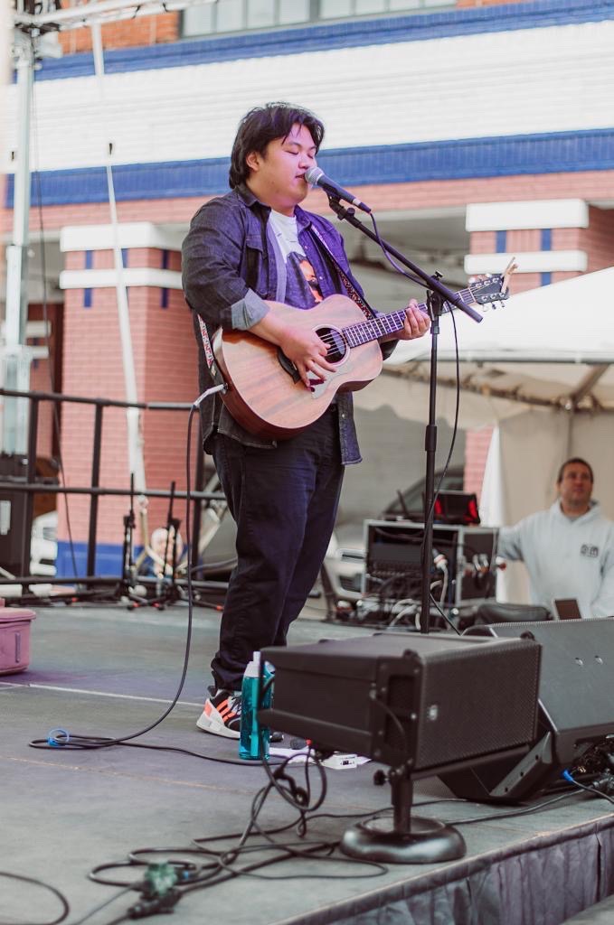 Person playing guitar and singing into microphone on stage outdoors, with a building and a person in the background.