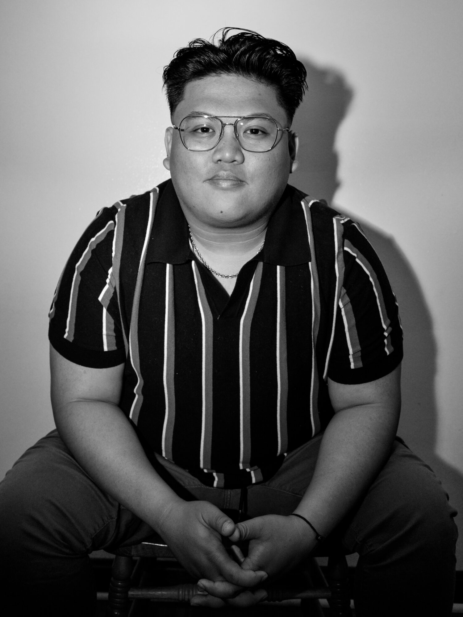 Young person with glasses and short hair, wearing a striped shirt, sitting with hands clasped, facing forward.