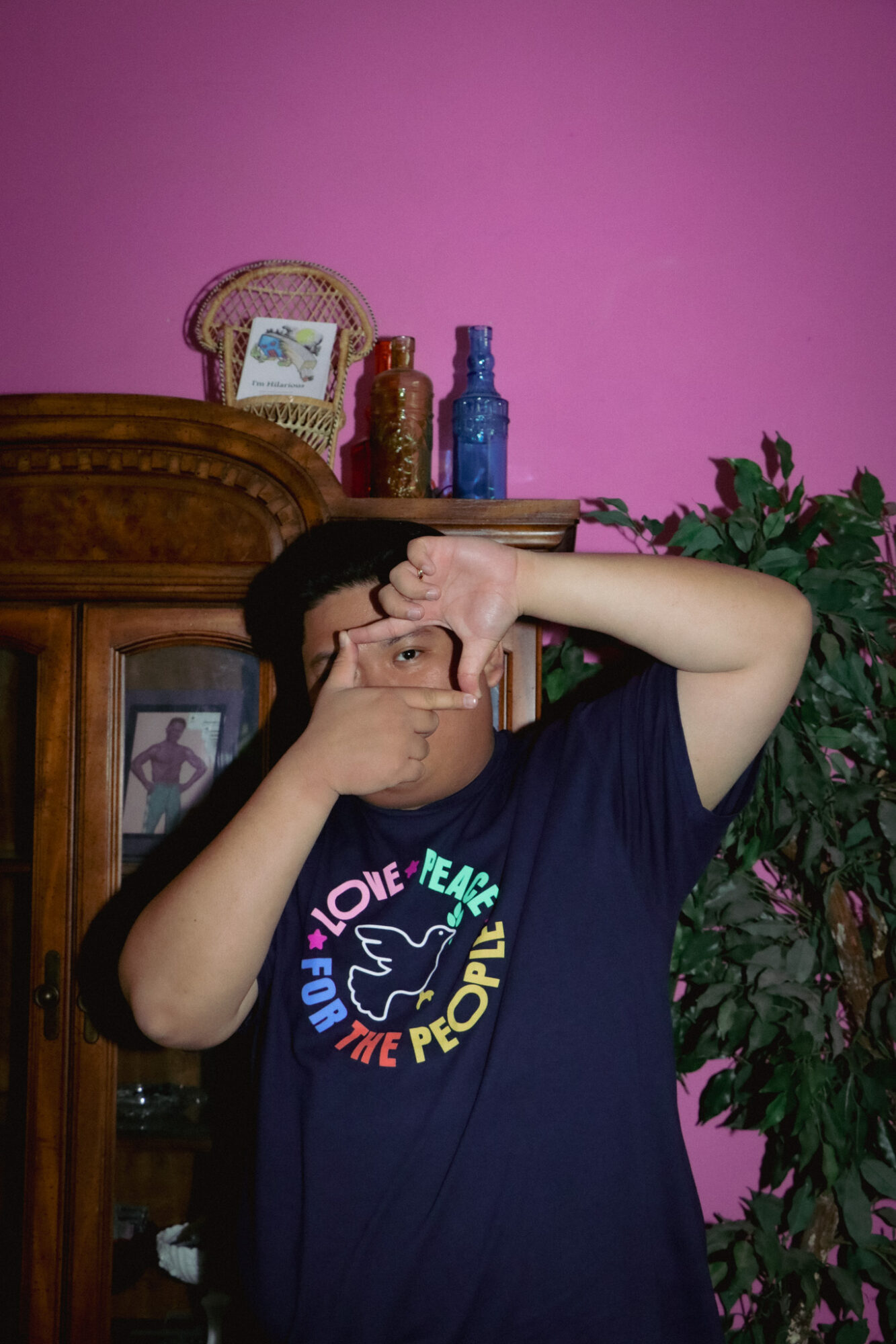 Person taking a selfie with a pink wall, wooden cabinet, and plants in the background.