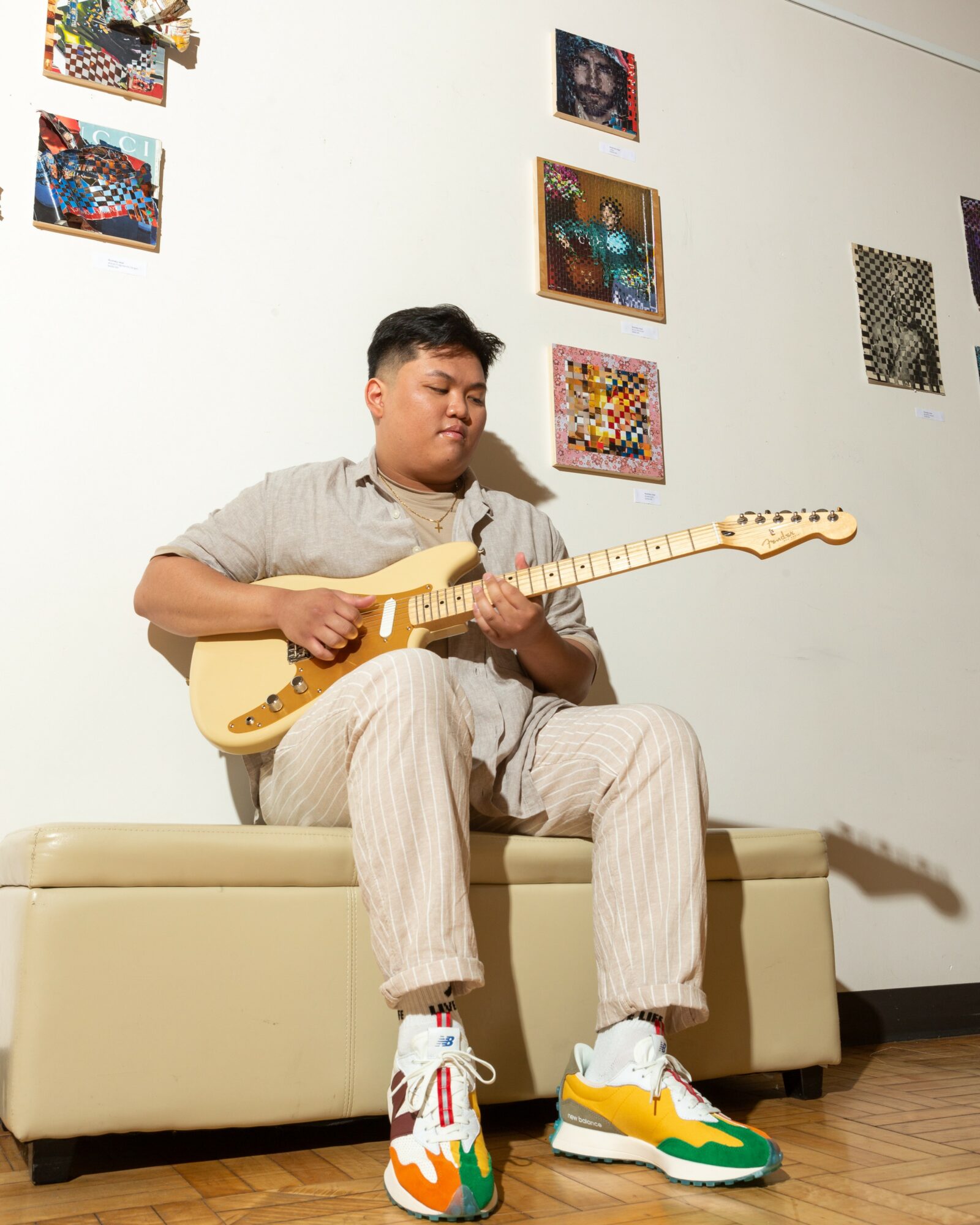Young man sitting on a beige bench playing an electric guitar, with colorful artwork on the white wall behind him.
