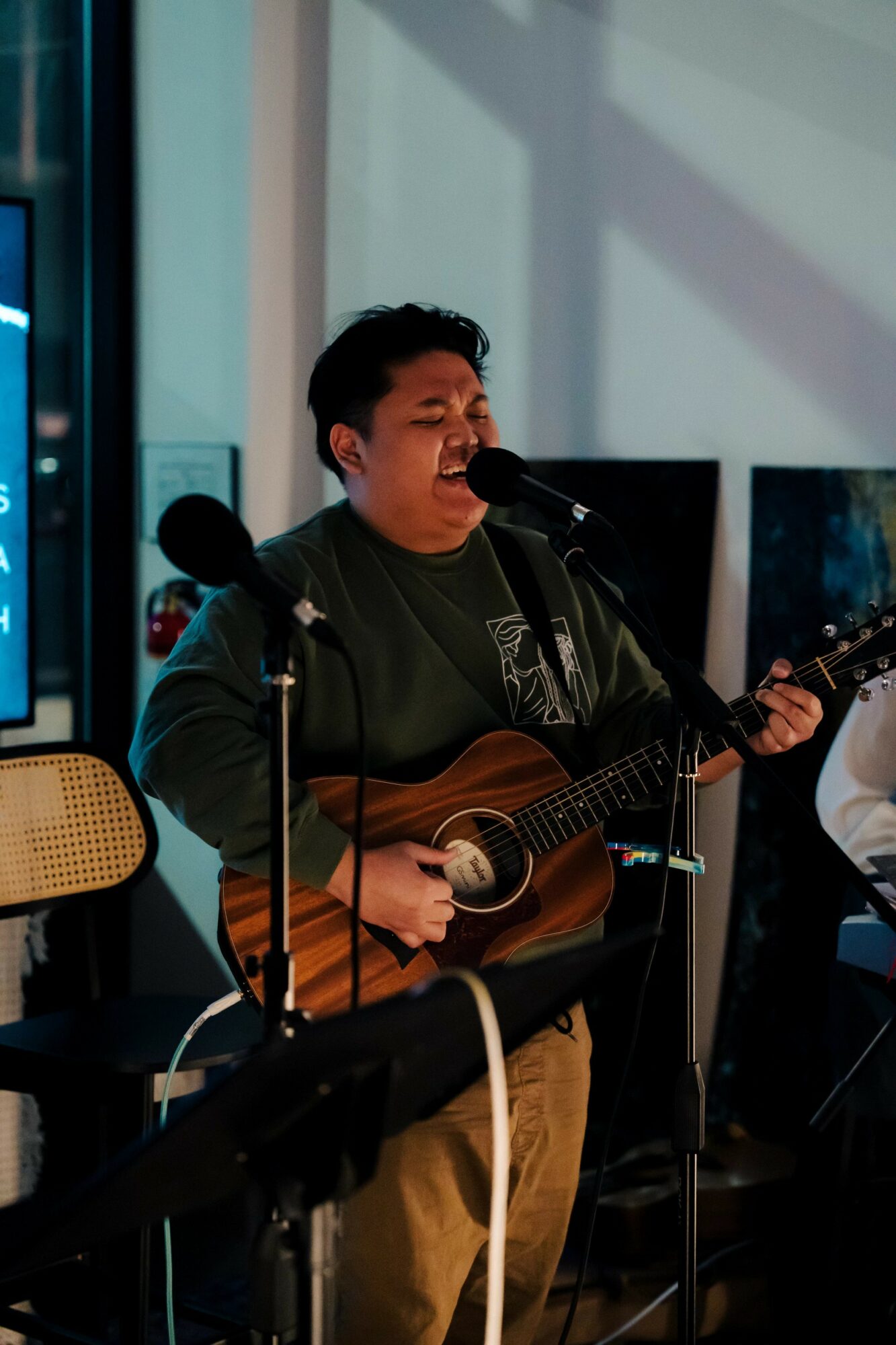 Person singing and playing guitar with microphones, in a room with large windows and electronic equipment.