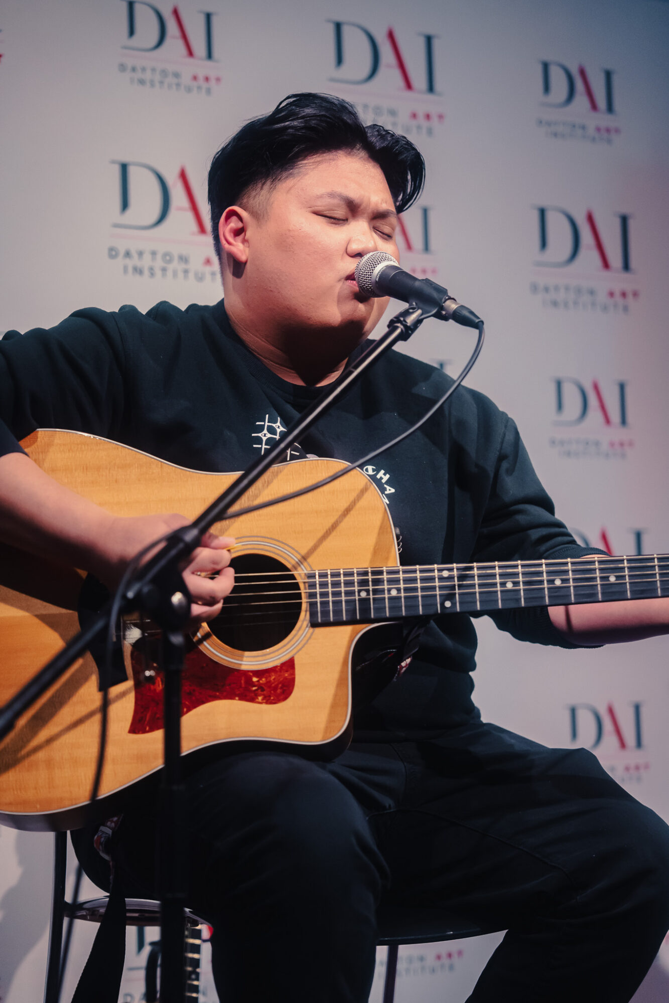 Person playing guitar and singing into microphone with a backdrop displaying 'D' and 'A' logos.