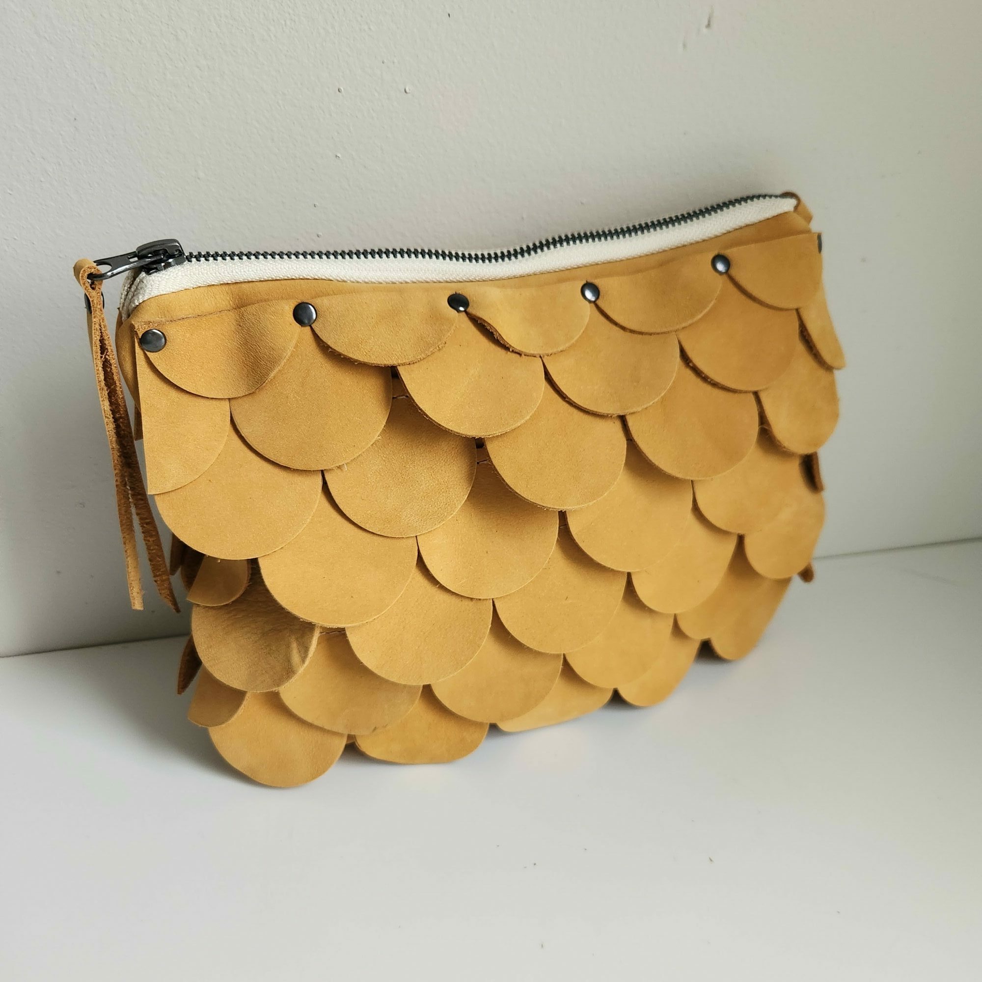 A tan clutch purse with a scalloped pattern and a zipper on the side, placed on a white surface.