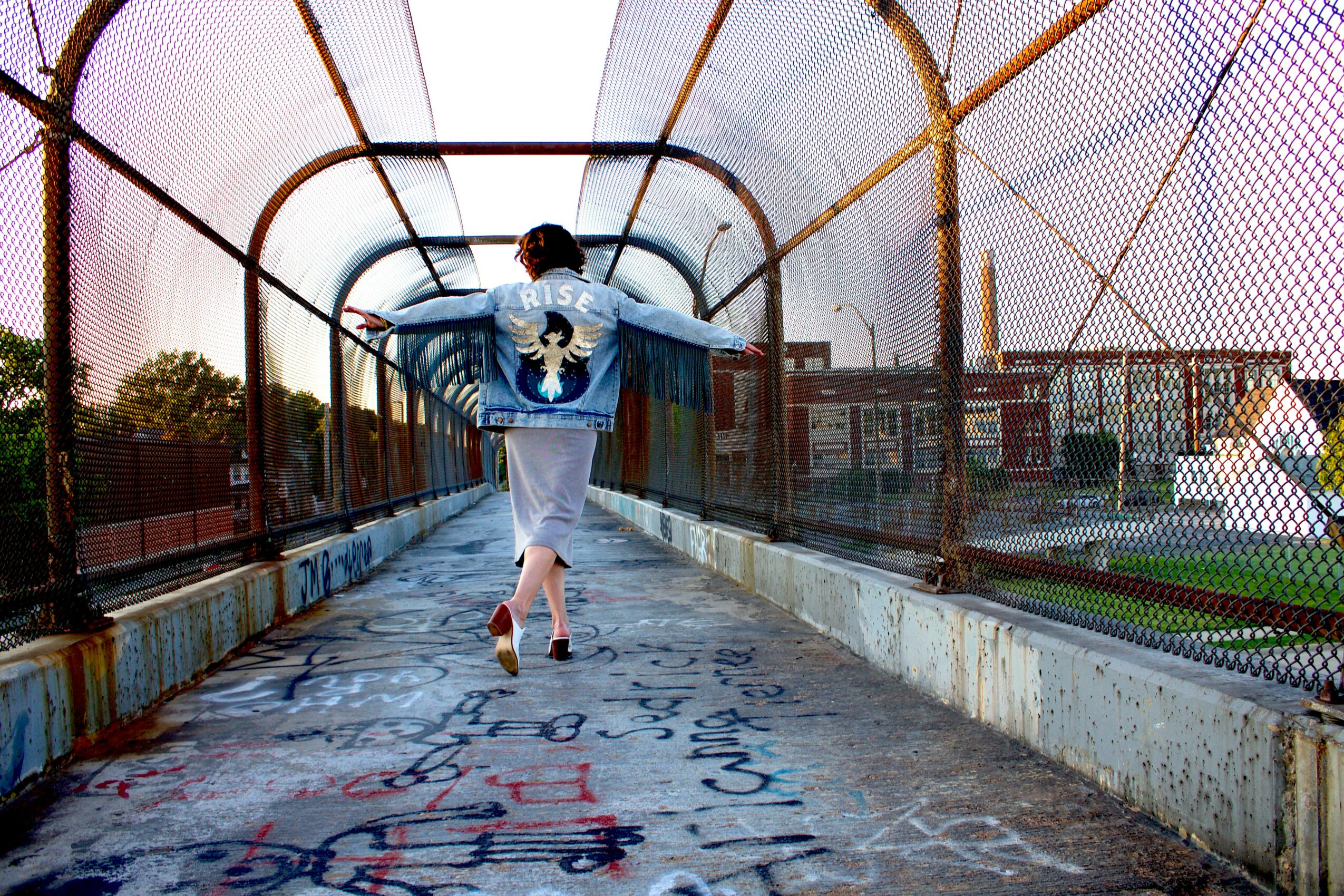 Person walking on a covered pedestrian bridge with colorful graffiti on the ground, metal railings, and cityscape in the background.