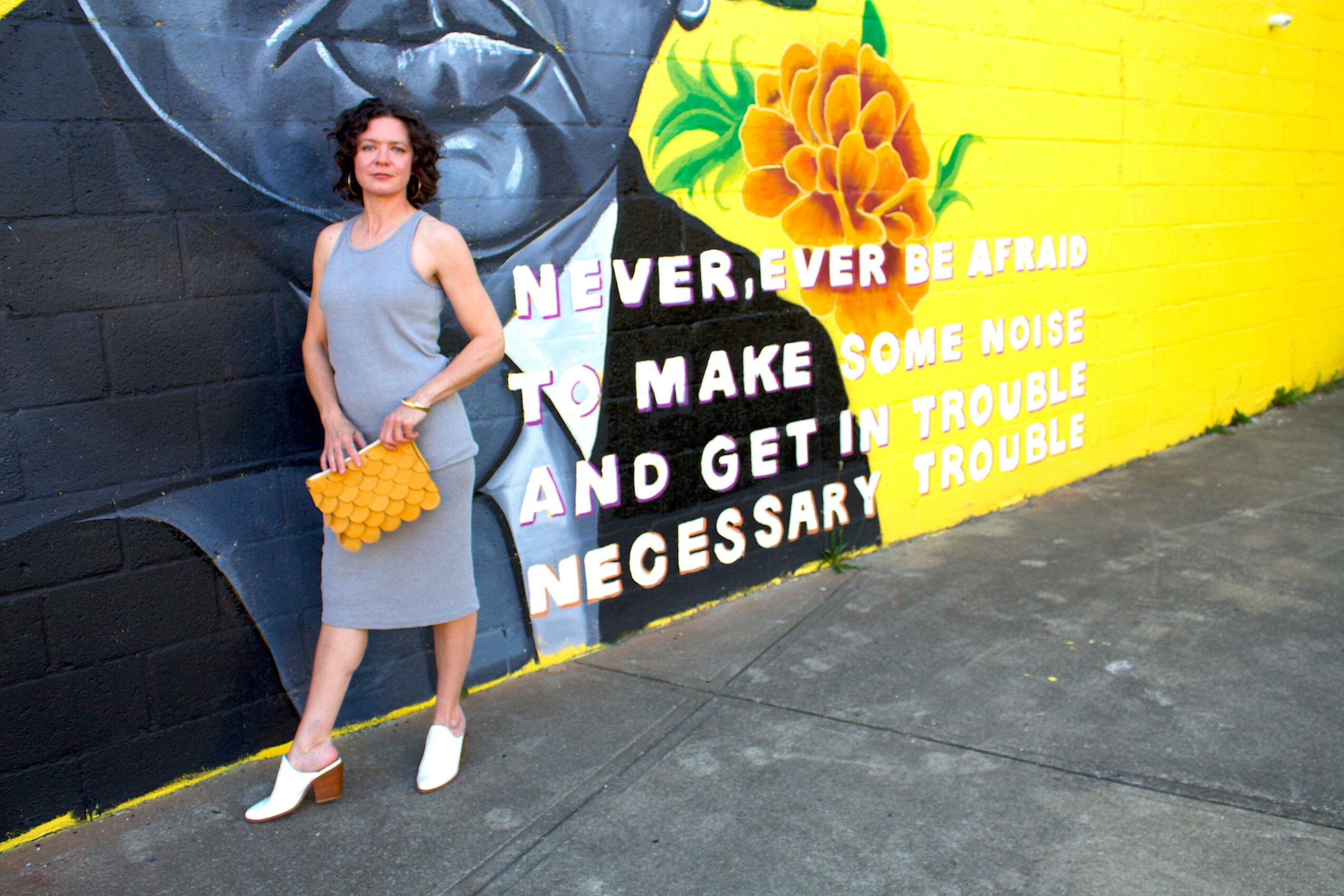 Woman in gray dress holding a yellow bag standing in front of a colorful mural with text and flowers, on a sidewalk.