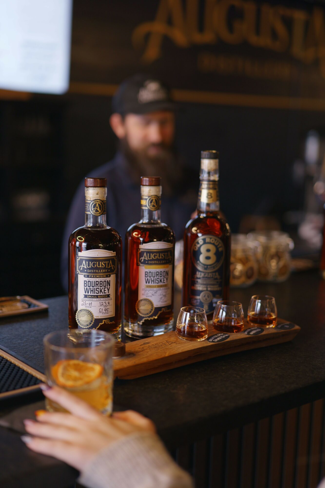 A wooden tray with several bottles of alcohol and a glass of whiskey on a dark table, with a person's hand holding a glass.