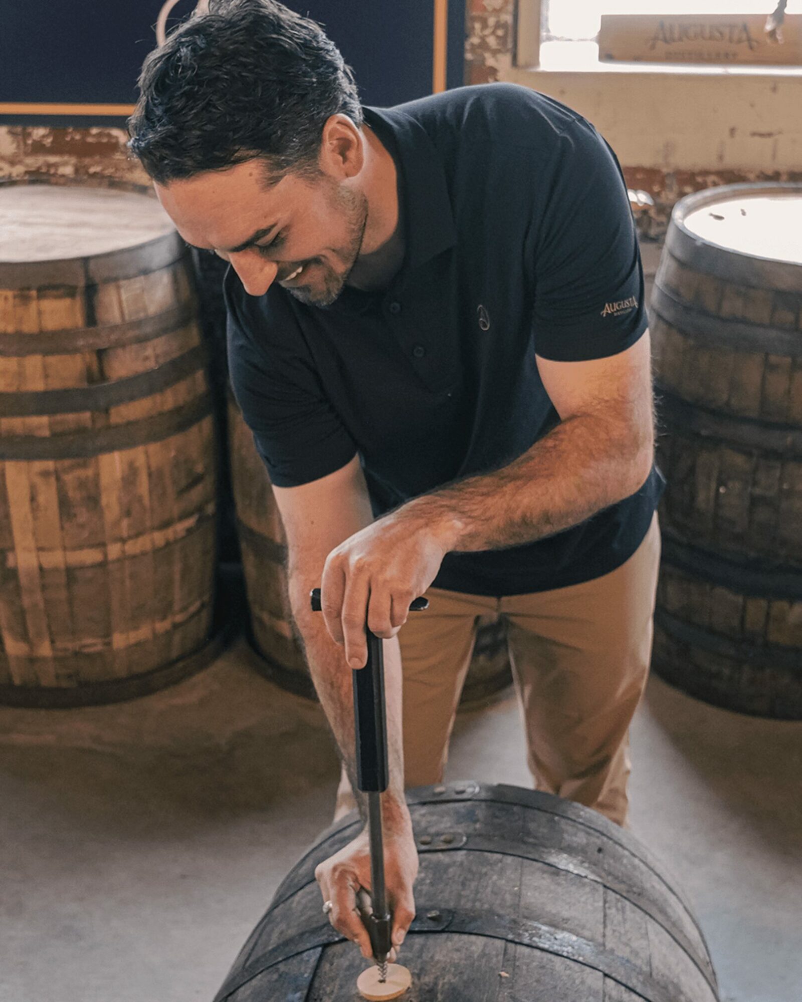 Man smiling and using a tool on a barrel in a room with wooden barrels and a window.