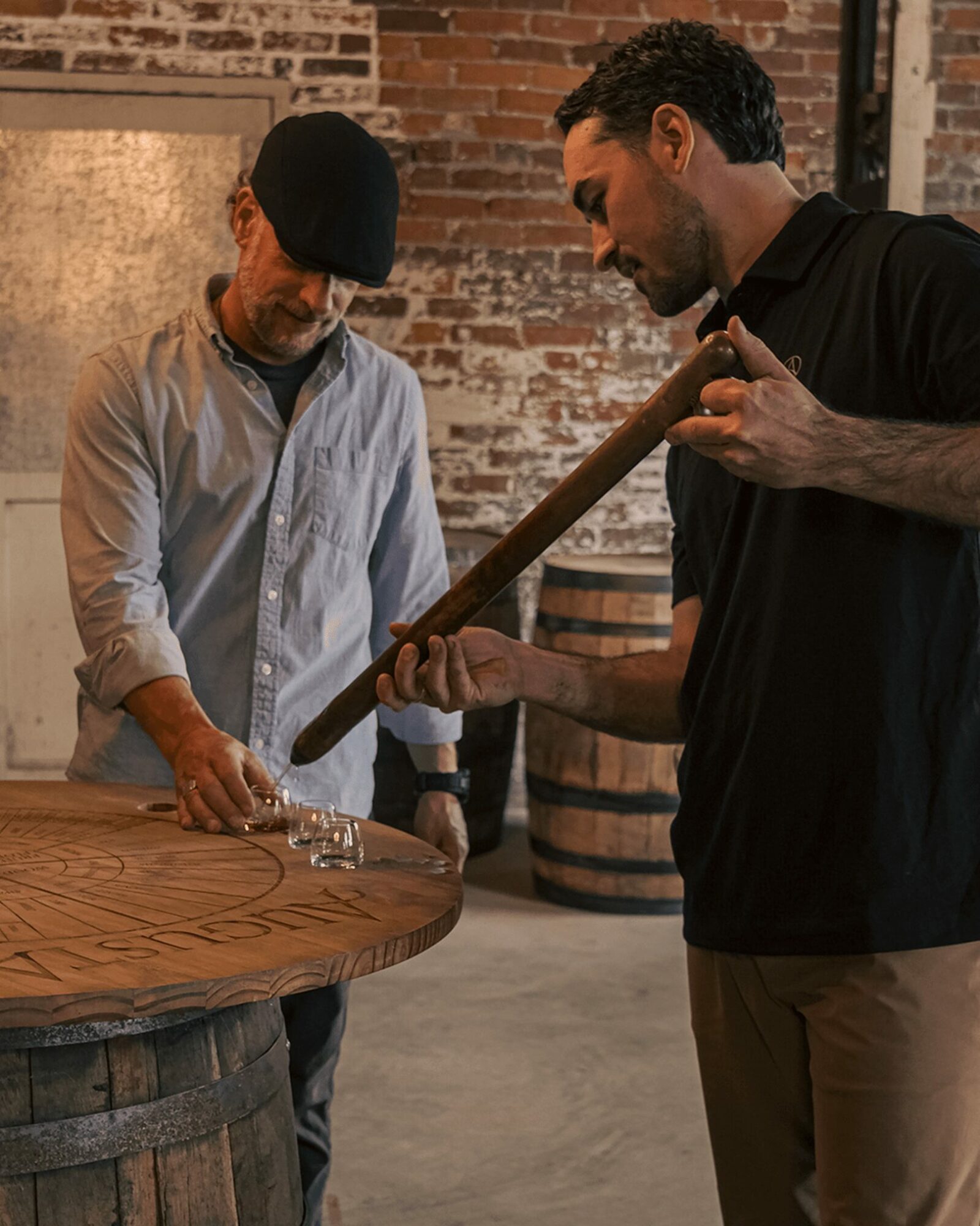 Two men are playing a game with a wooden stick and small glass pieces on a round table in a brick-walled room.