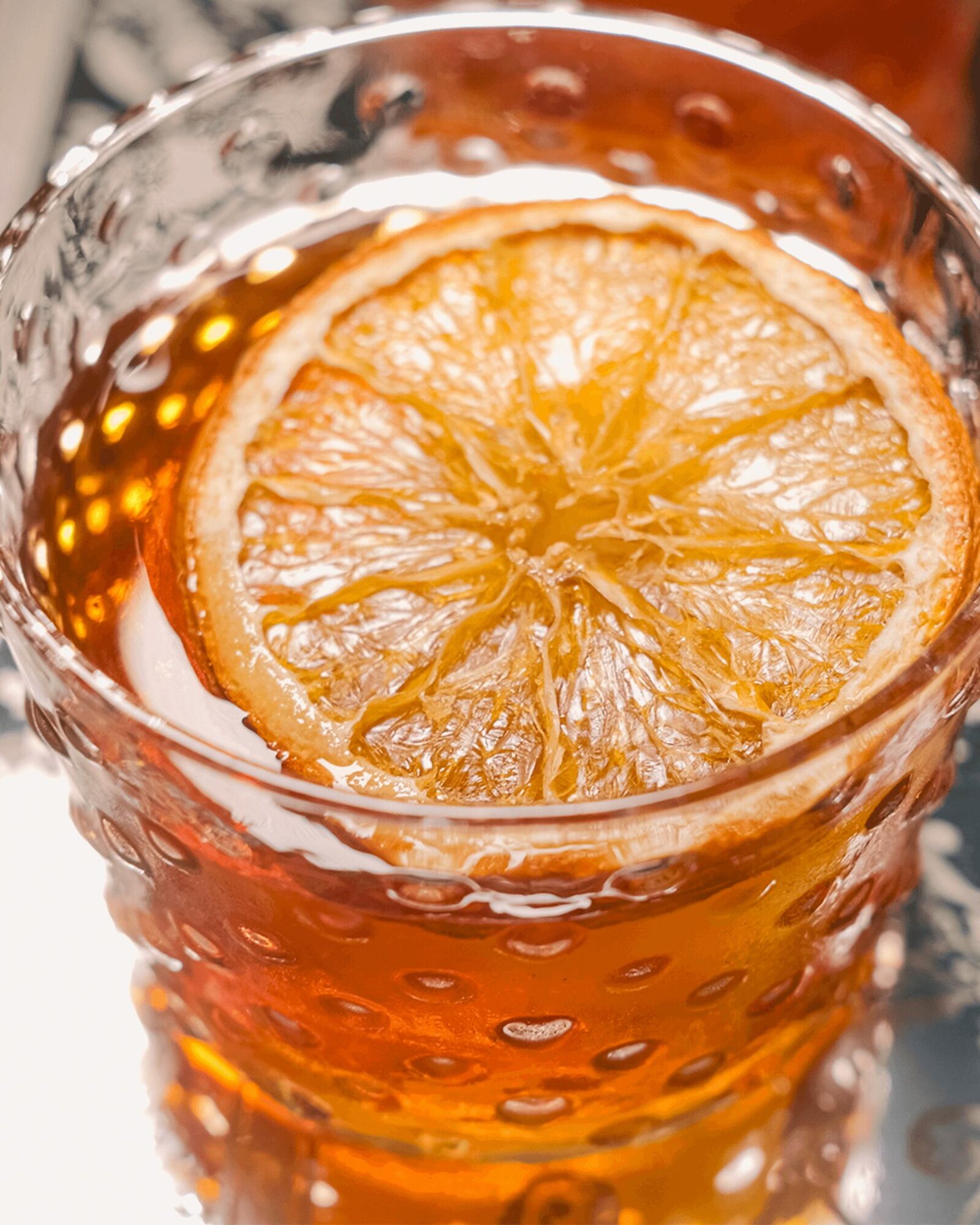 A glass with a slice of dried lemon on top, filled with a dark beverage, against a blurred background.