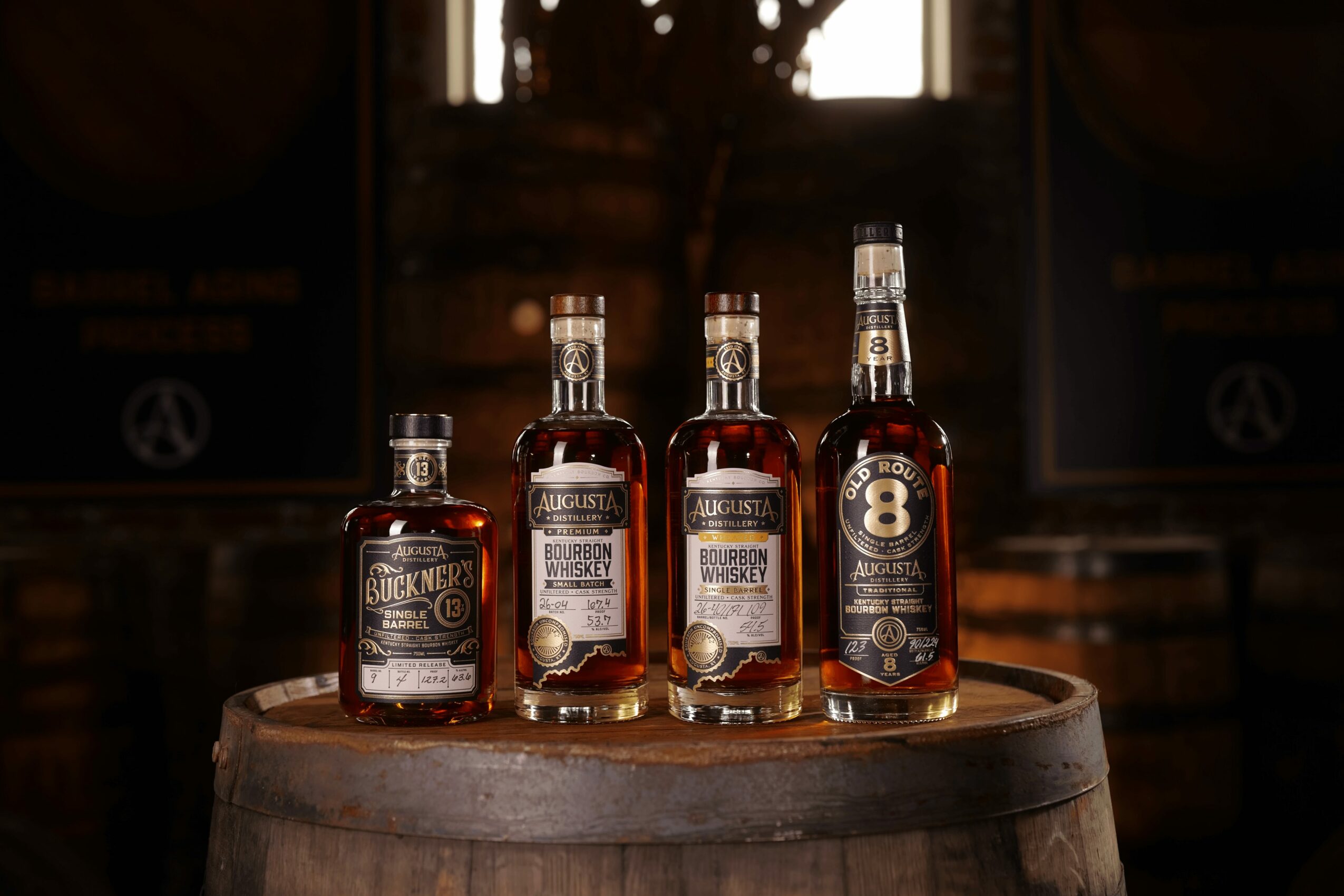 Four bottles of whiskey on a wooden barrel in a dimly lit setting, with barrels in the background.