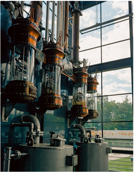 Industrial equipment with glass containers and pipes inside a building with large windows, natural light, and trees outside.
