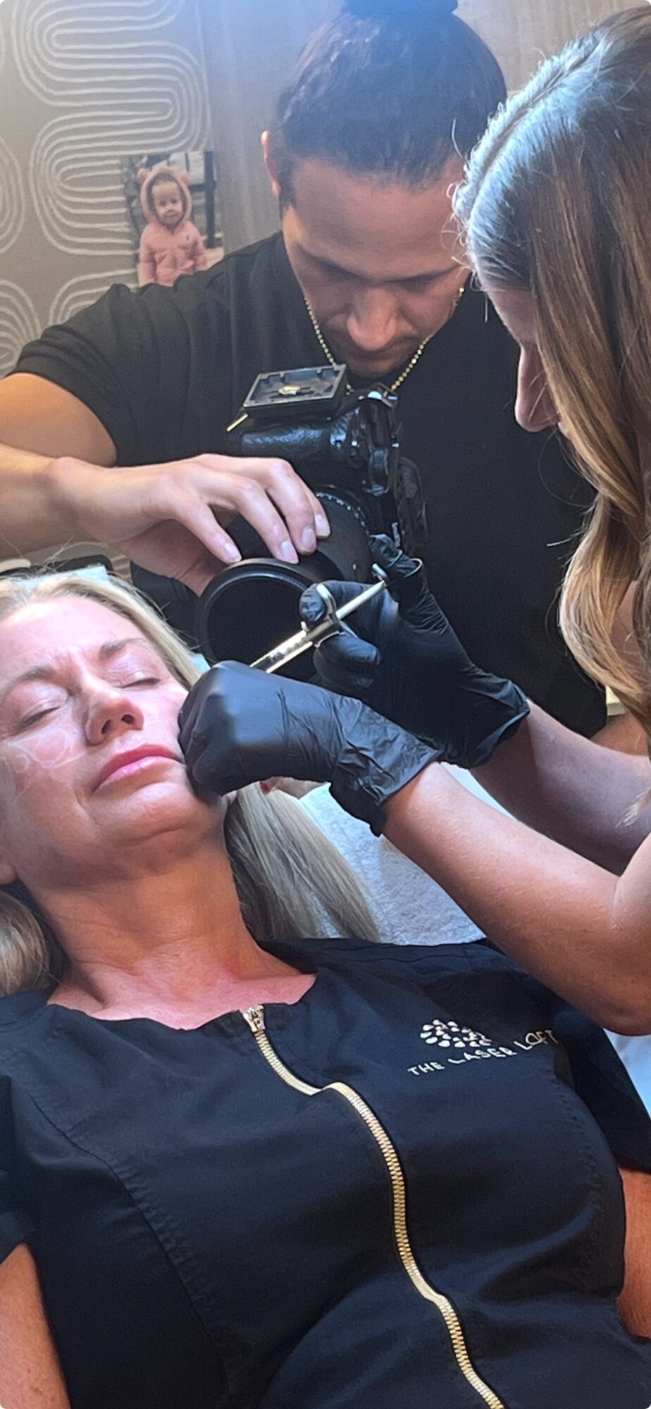 Woman receiving a cosmetic procedure on her face from a professional, with a photographer capturing the moment.