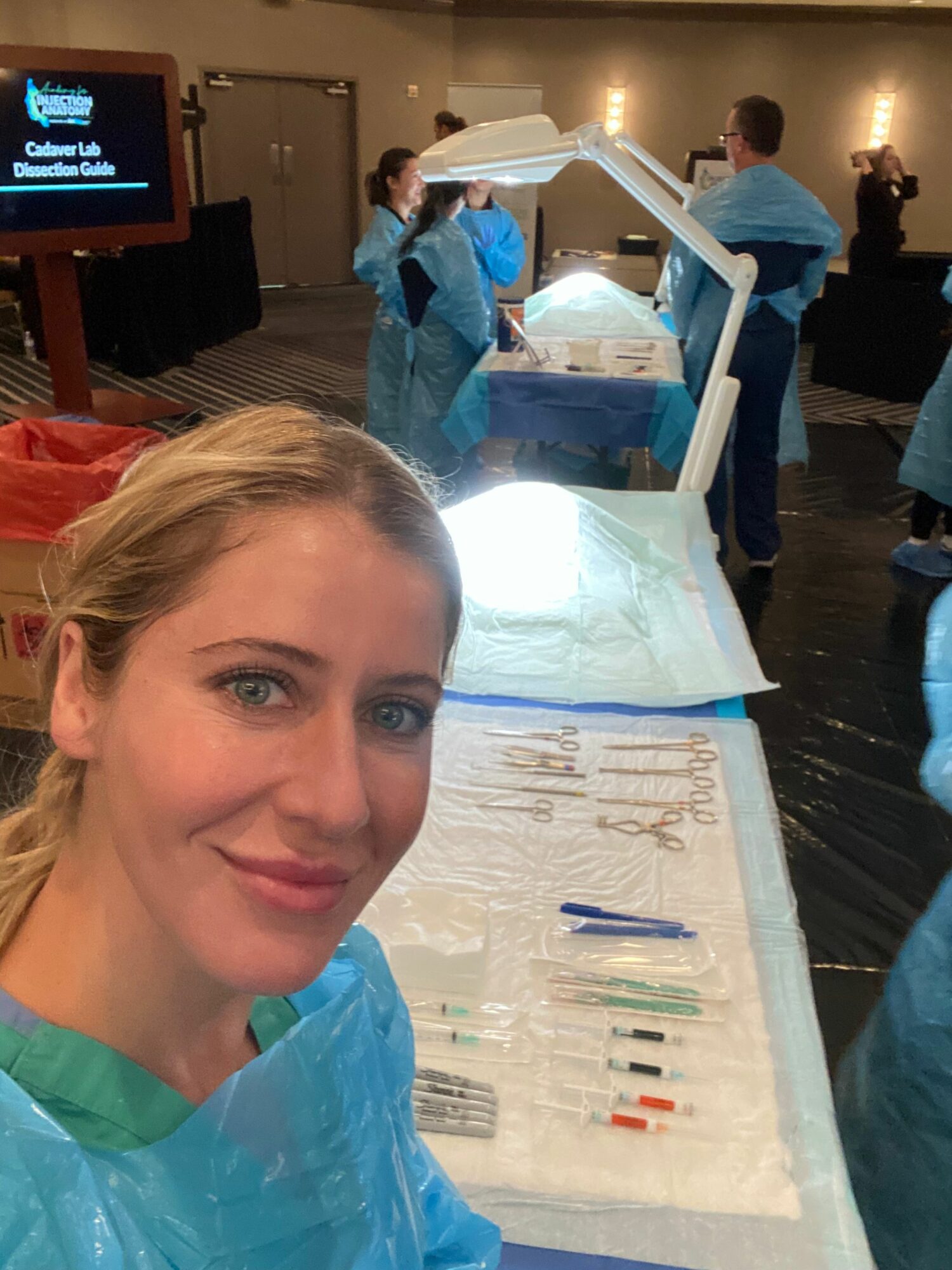 Woman with blonde hair in a blue protective gown, lying on a medical bed with medical tools nearby, in a clinical setting.