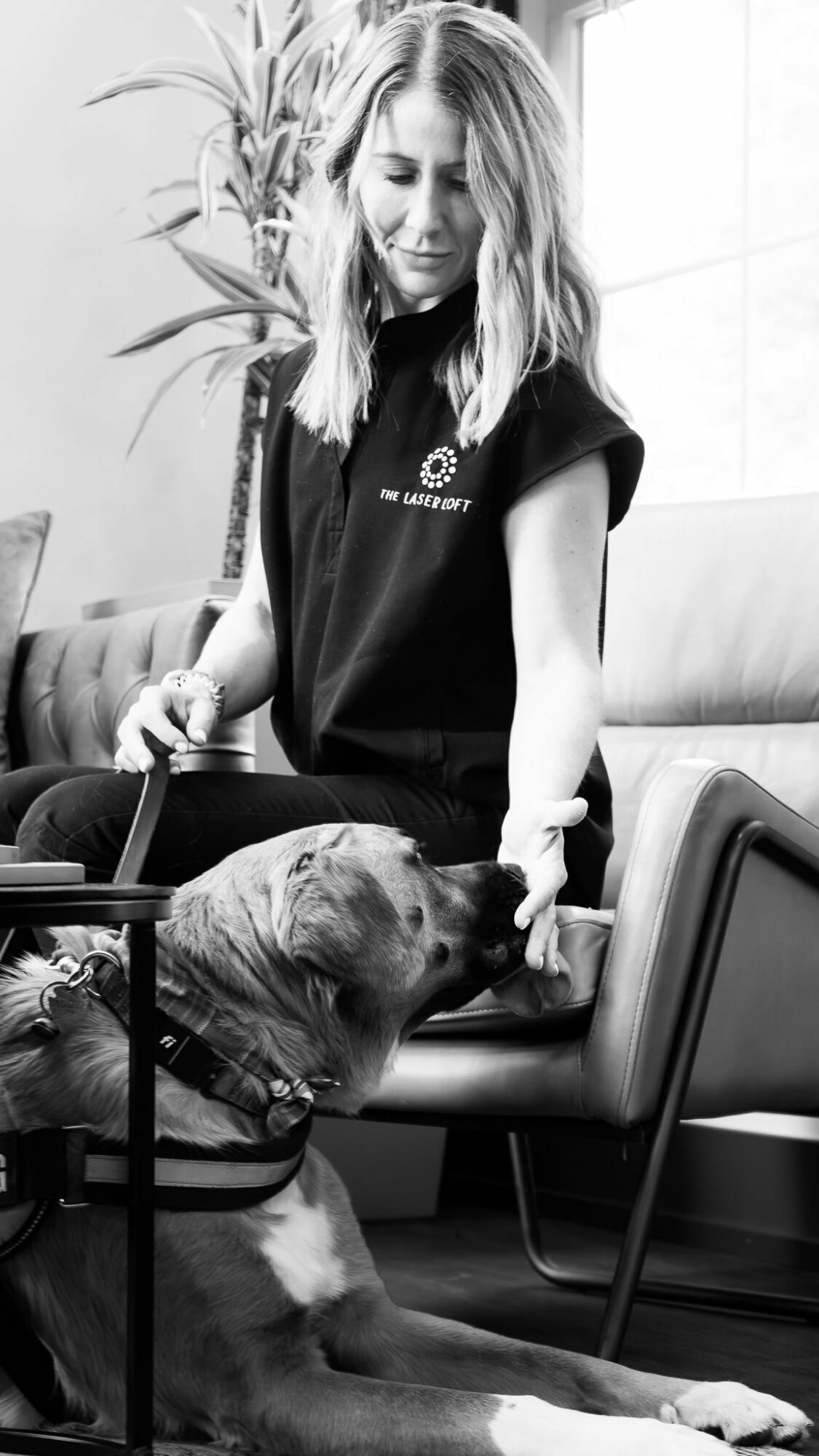Young woman with long hair interacts with a dog sitting on a chair in a room with a plant in the background.
