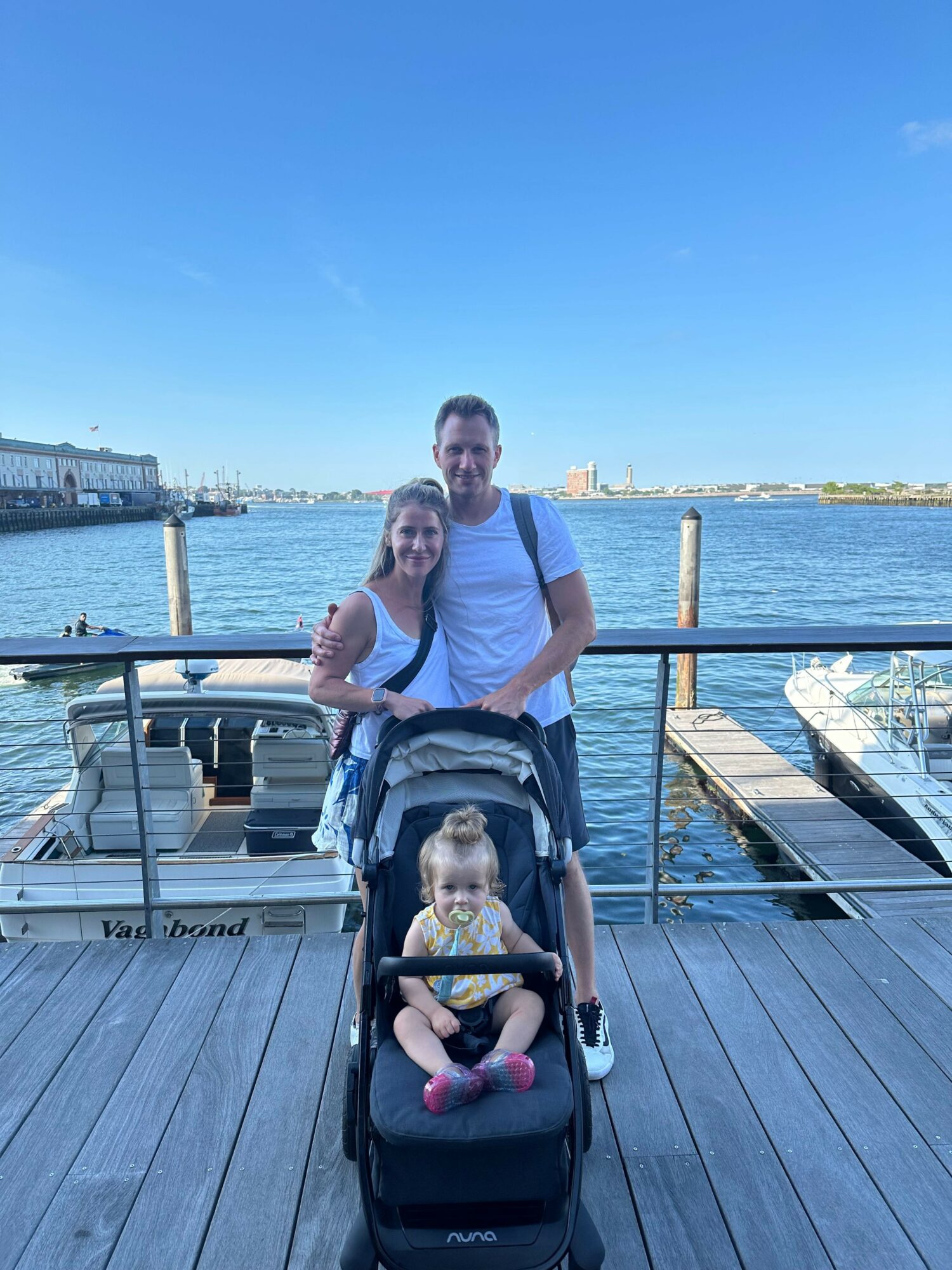 Two people stand on a wooden dock by water, with a stroller and a child, under a clear blue sky.