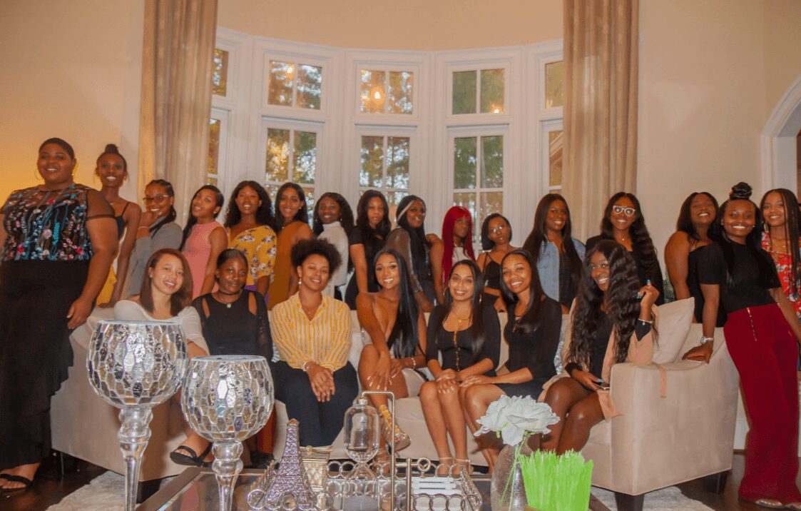 Group of women and girls gathered in a living room with large windows, some sitting and some standing, smiling.