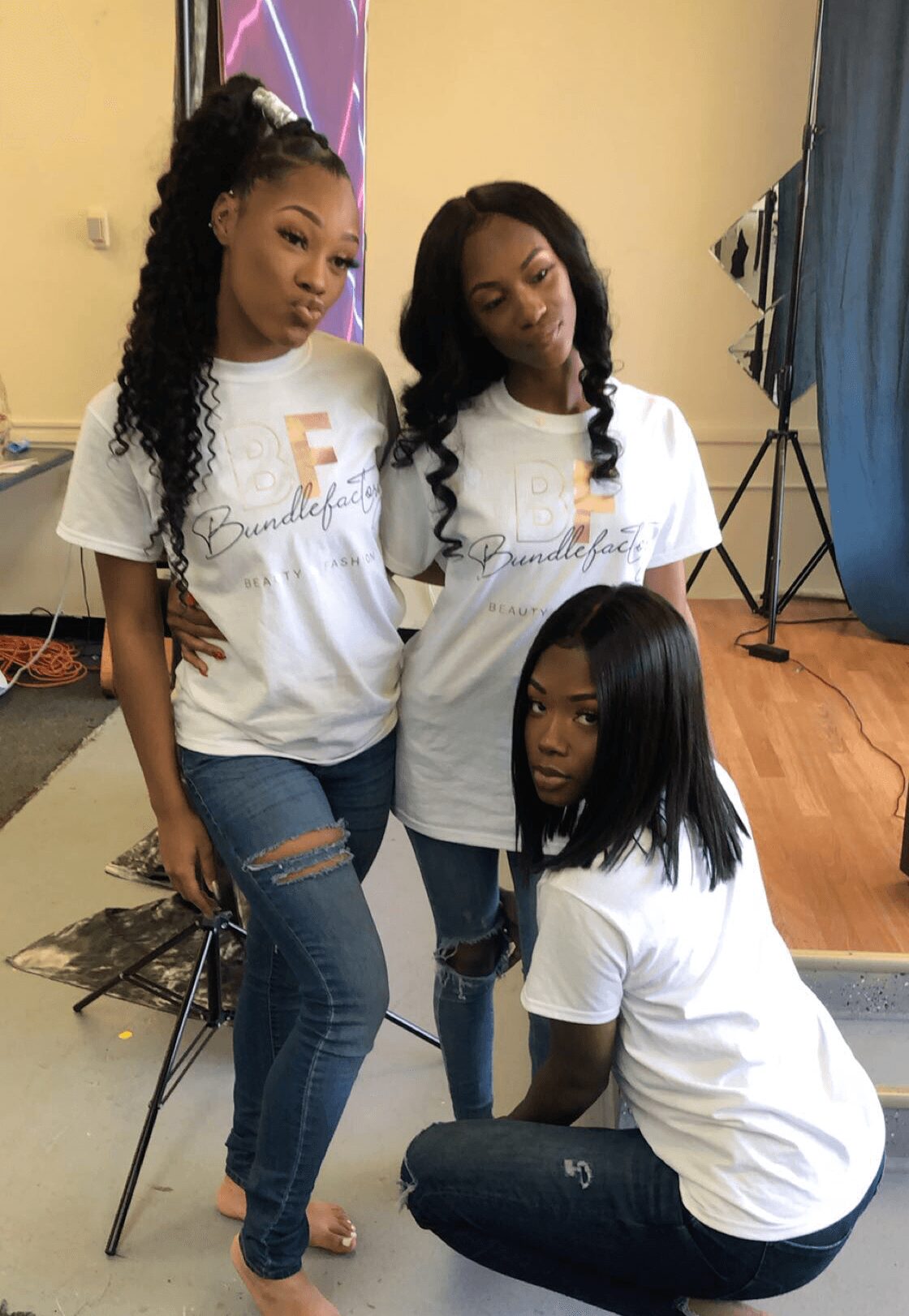 Three women in white T-shirts and jeans pose indoors, two standing and one kneeling, with a yellow wall and blue curtain in background.