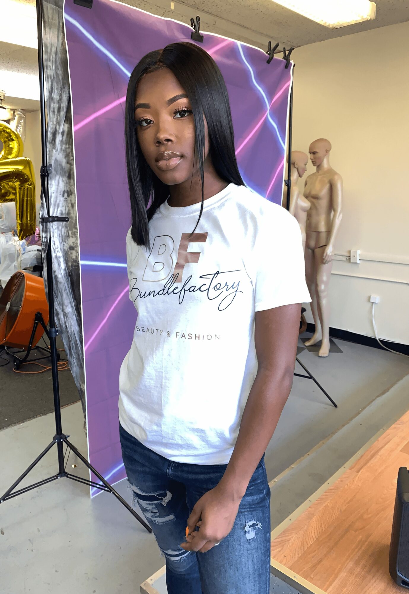 Young woman with straight black hair wearing a white T-shirt and ripped jeans, standing indoors with a mannequin and neon backdrop.