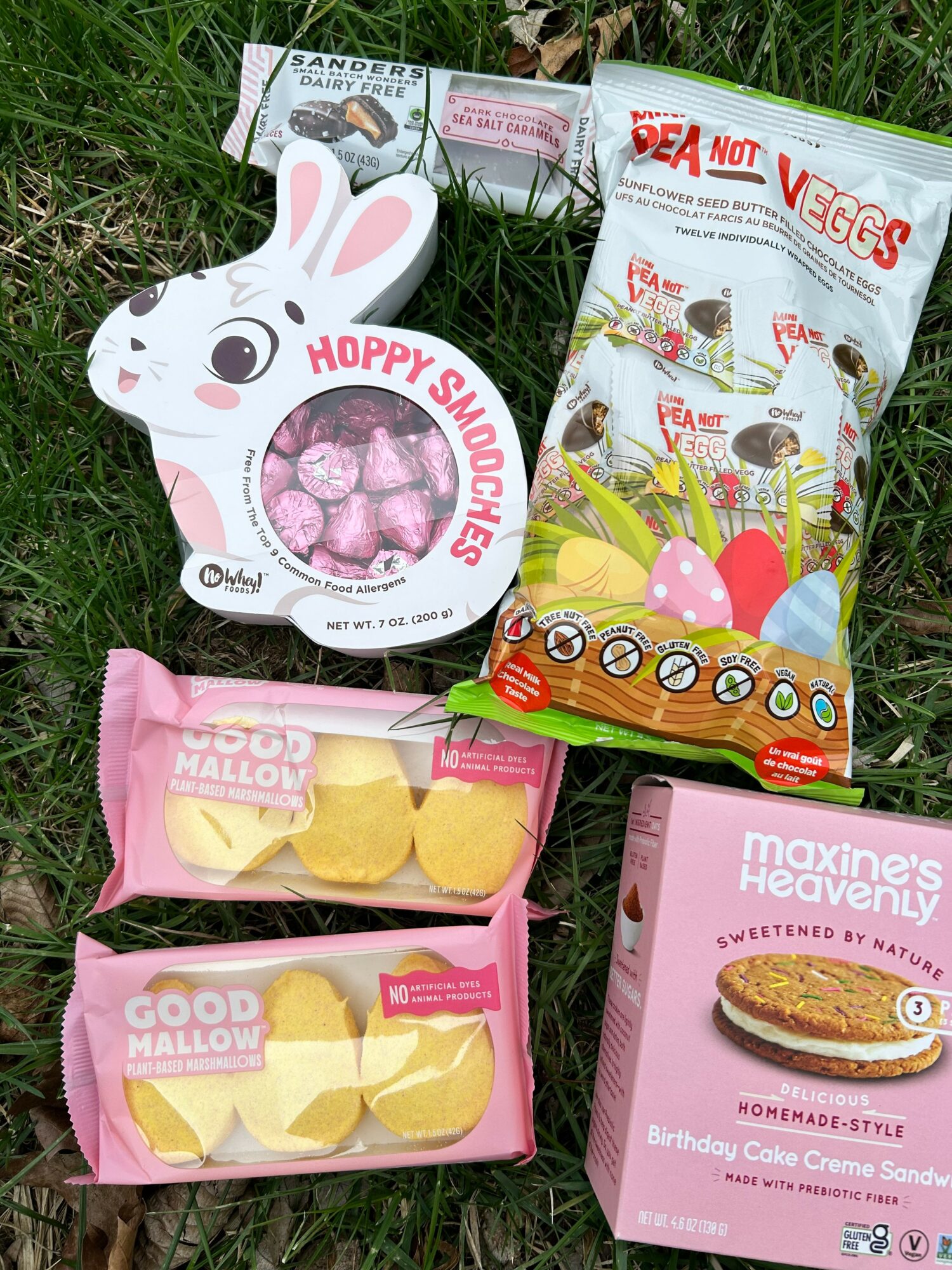 Assorted snack packages and a bunny-shaped container with pink candies on grass.