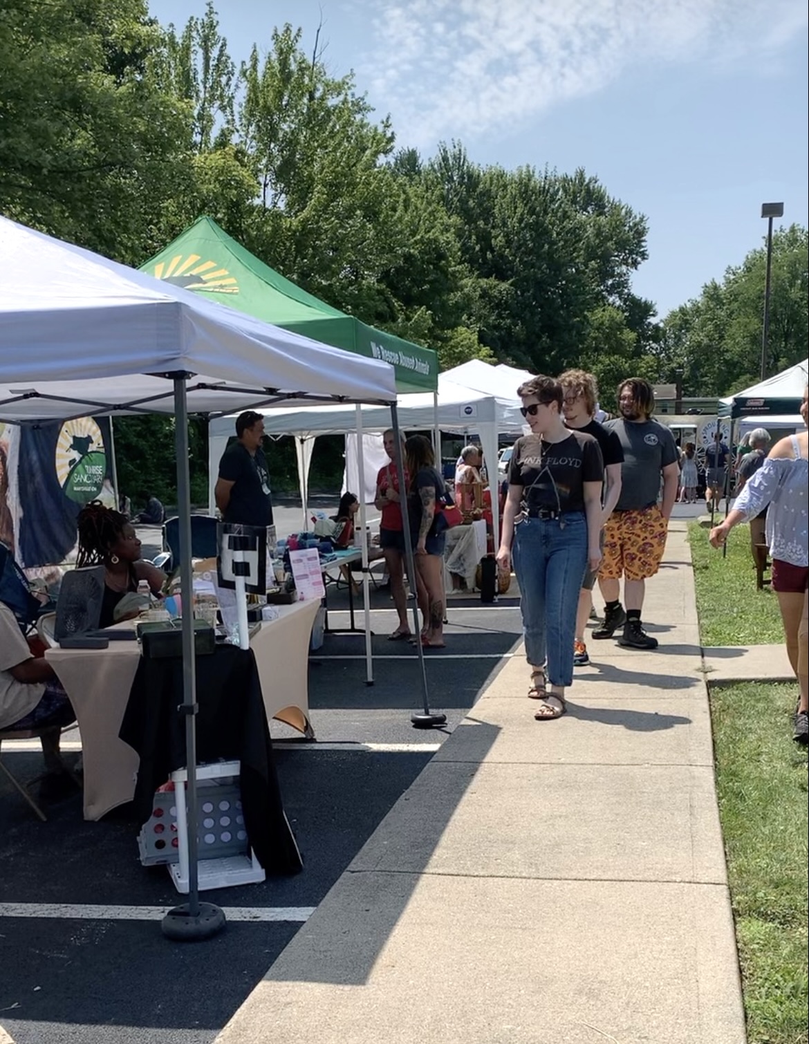 People walking along a sidewalk at an outdoor event with tents and trees in the background.