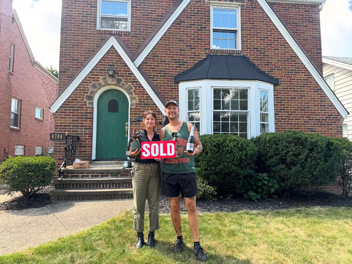 Two people standing in front of a brick house with a bay window and green bushes, holding a red 'SOLD' sign.