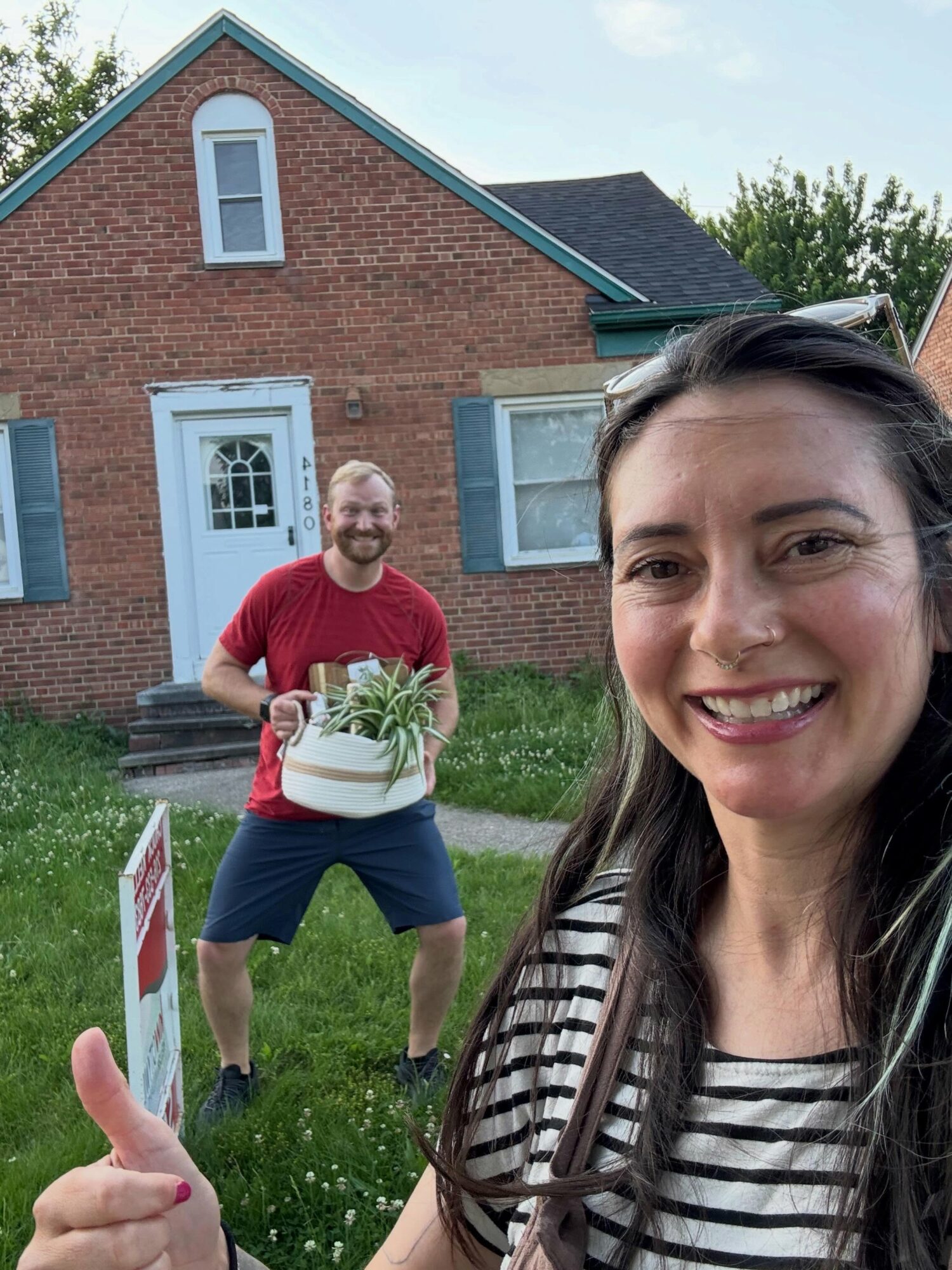 Two people outdoors in front of a brick house, smiling, with one person holding a potted plant, woman taking a selfie.