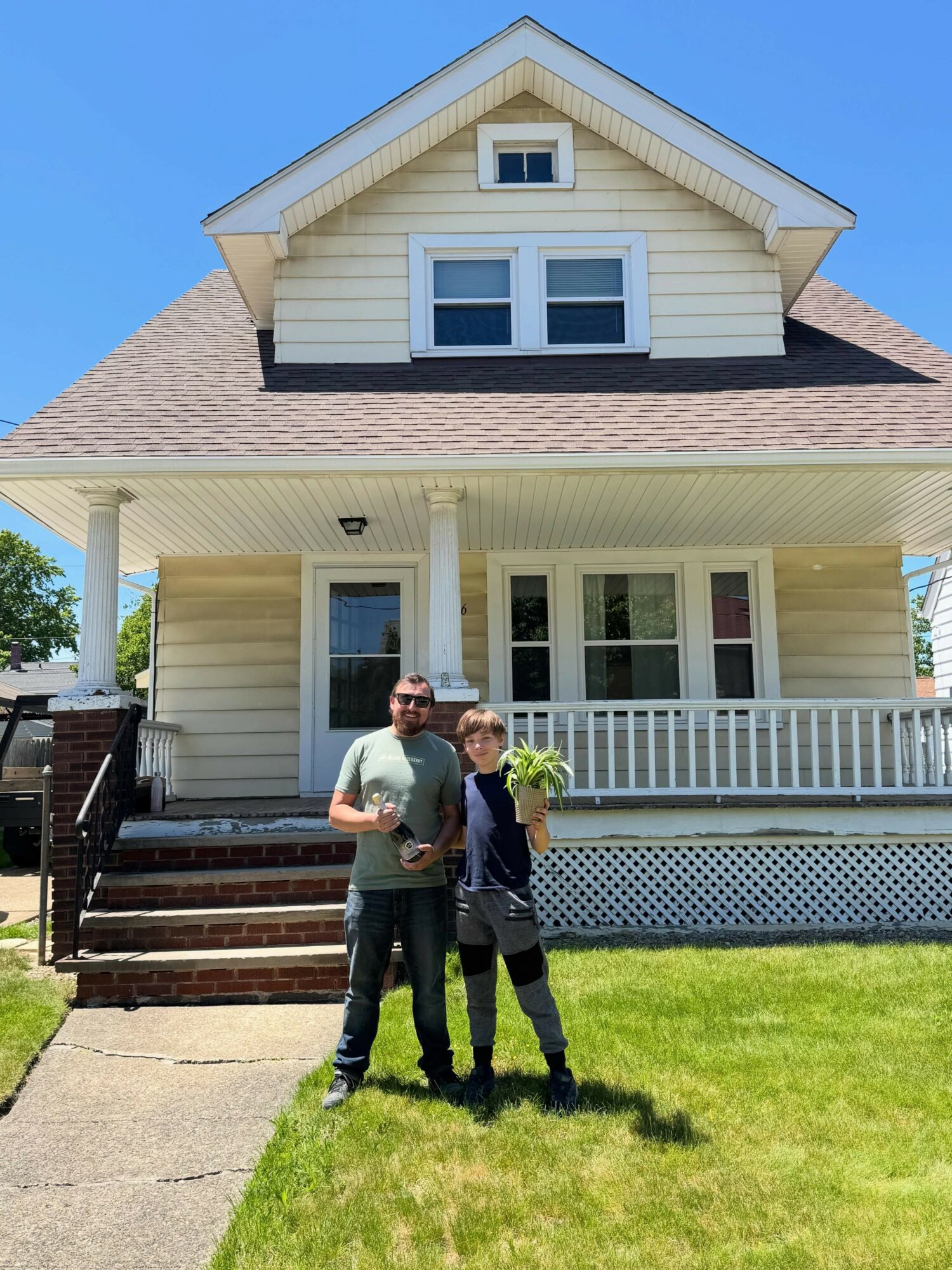 Three people standing on front lawn in front of a two-story house with porch, white siding, and gabled roof.