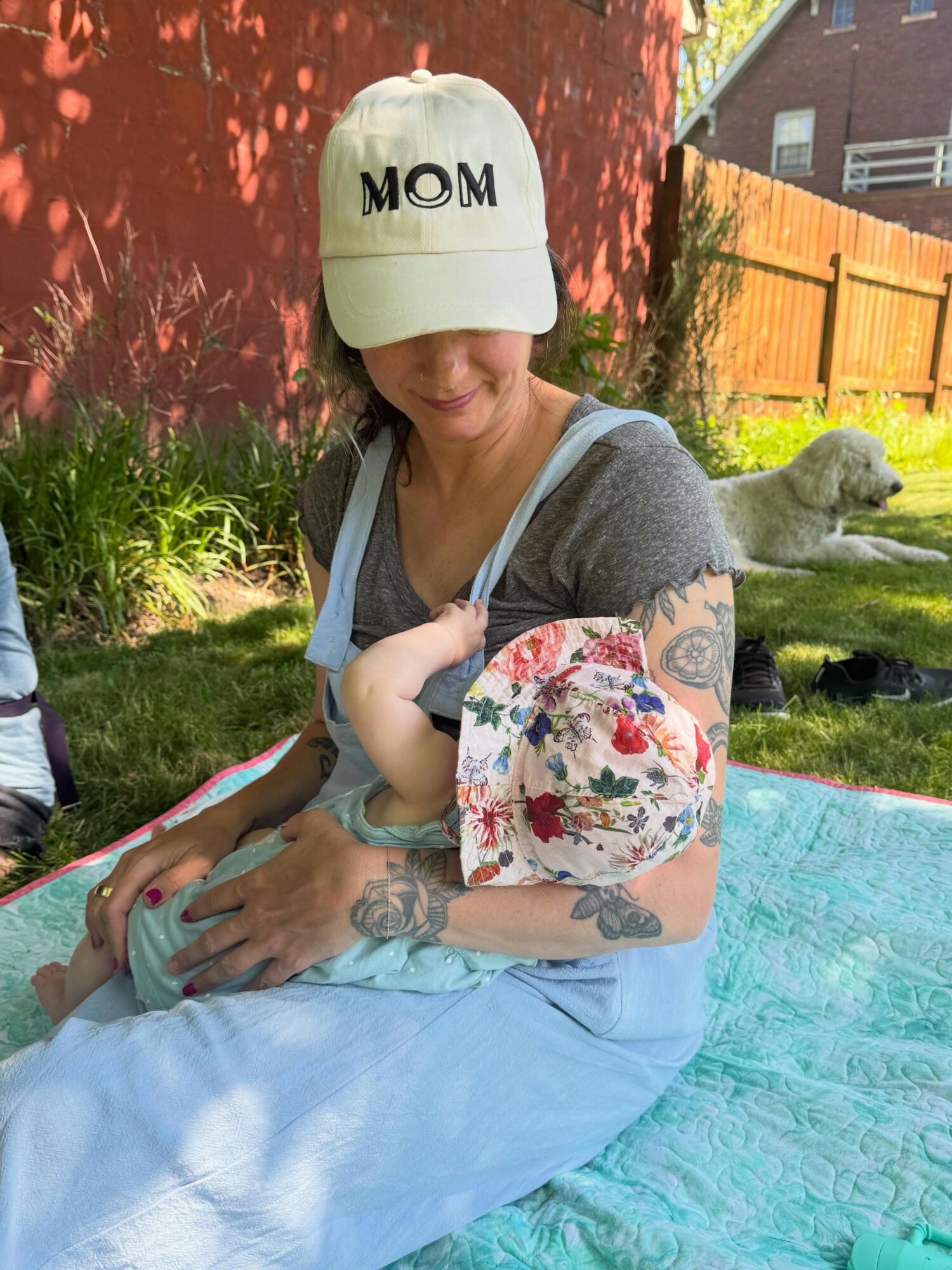 Woman wearing a cap with 'MOM' and holding a baby, sitting outdoors on a blanket, with a dog in the background.