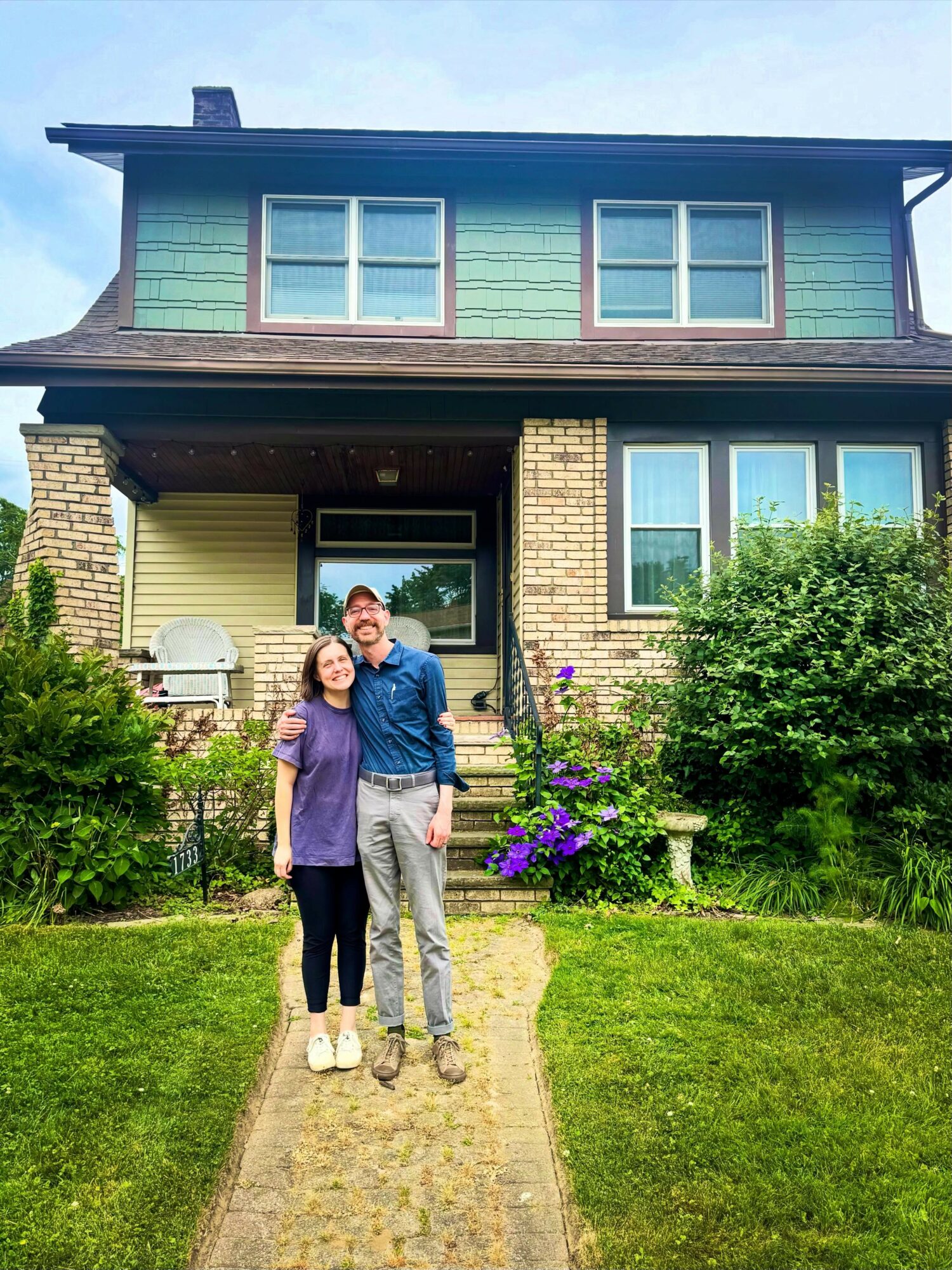 Two people stand on a stone pathway in front of a two-story house with green and yellow exterior, surrounded by greenery.