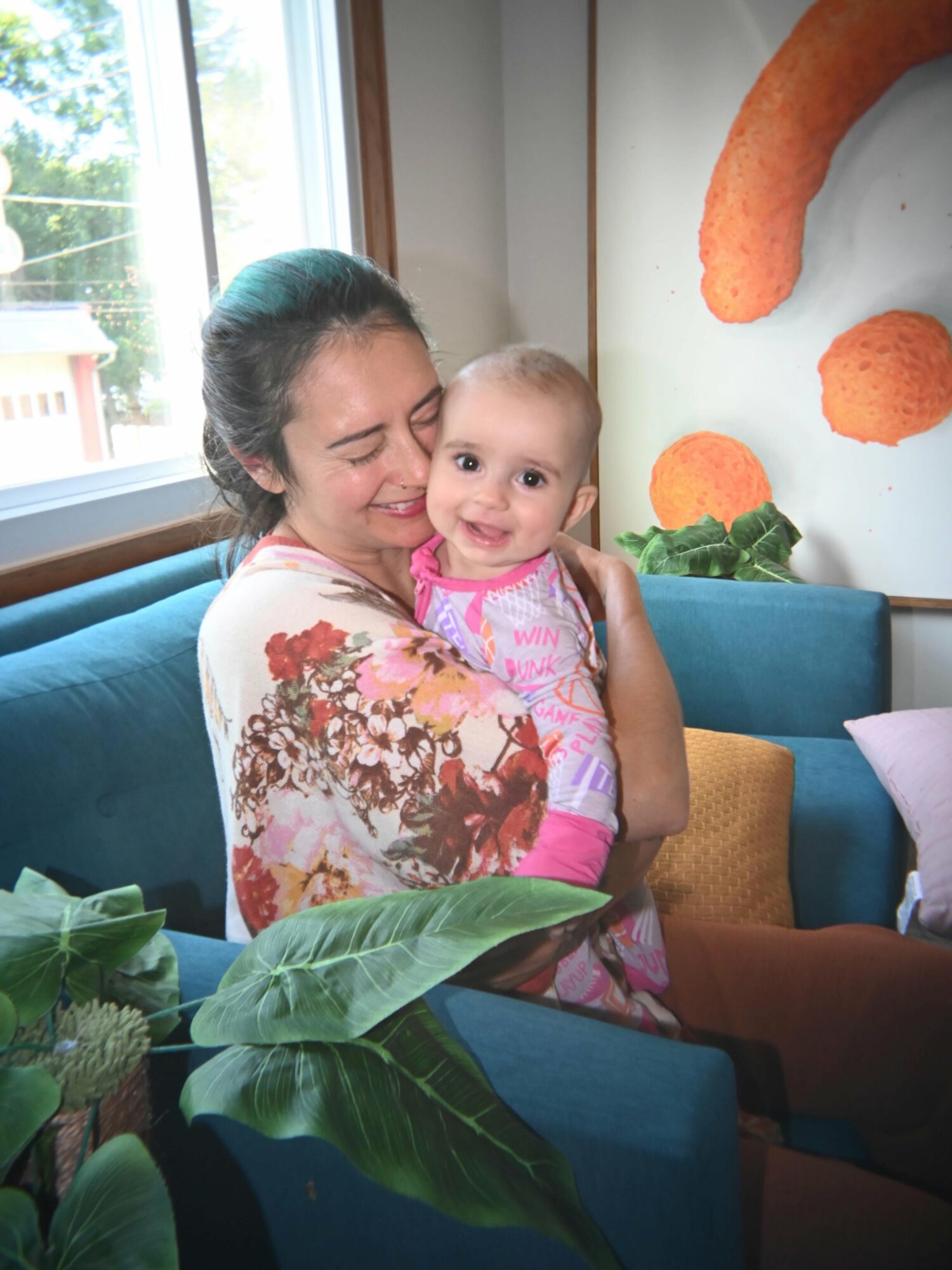 Woman holding smiling child indoors near window, plants and colorful wall art visible.