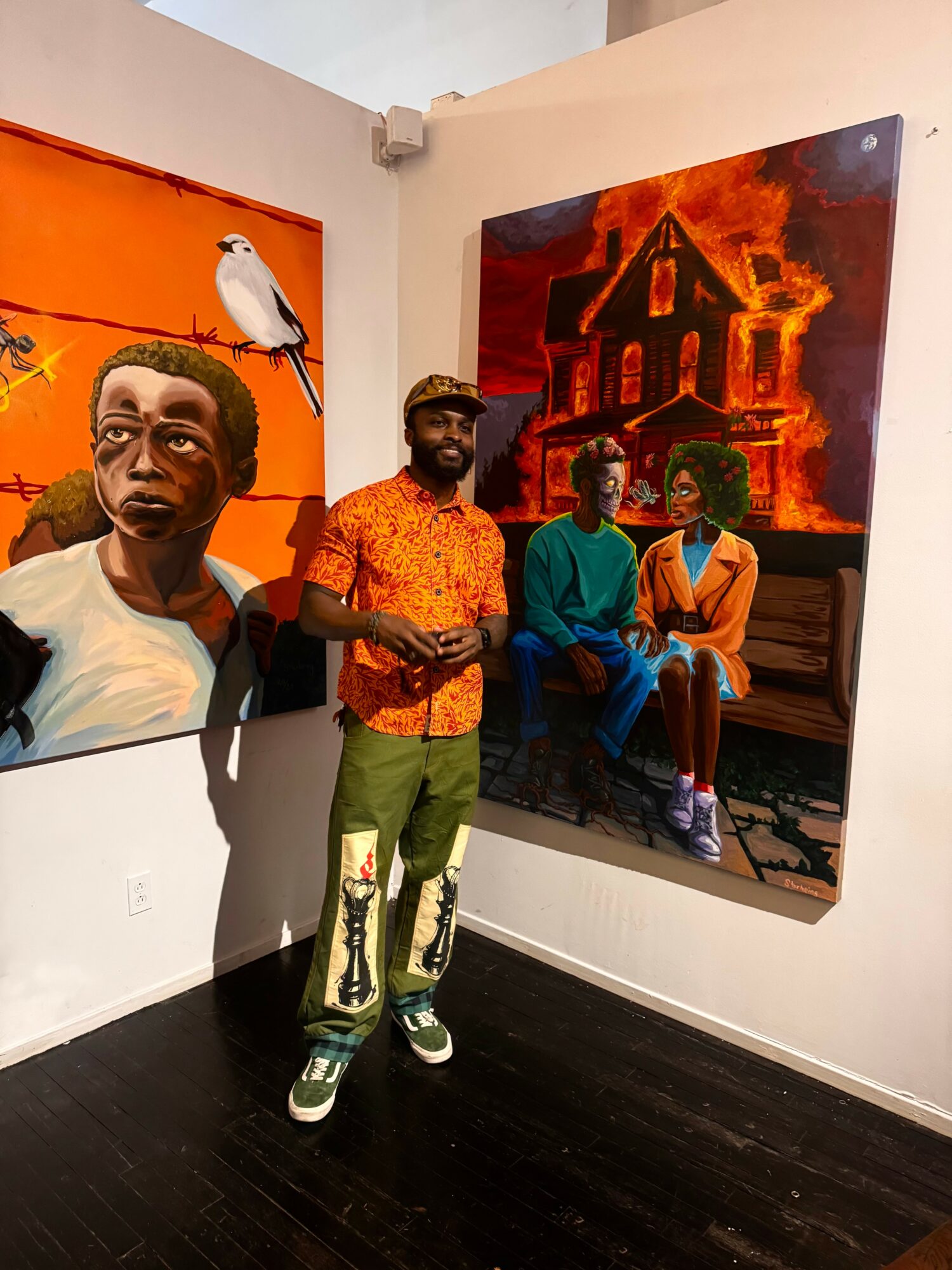 Man standing in front of two colorful paintings, one with a woman and bird, the other with people and a house on fire.