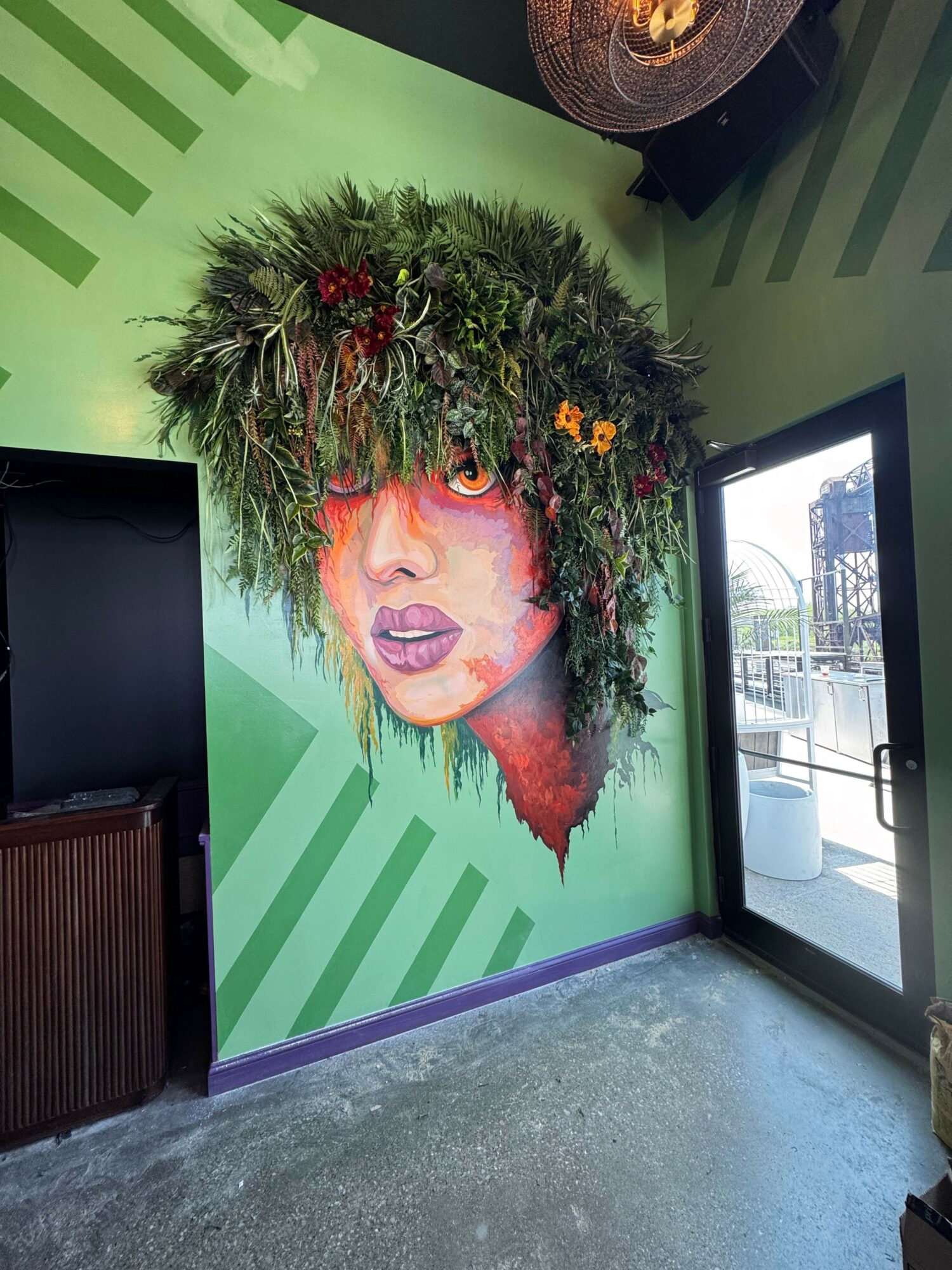 Wall mural of a woman's face with a headdress made of greenery and flowers, on a green background with stripes.