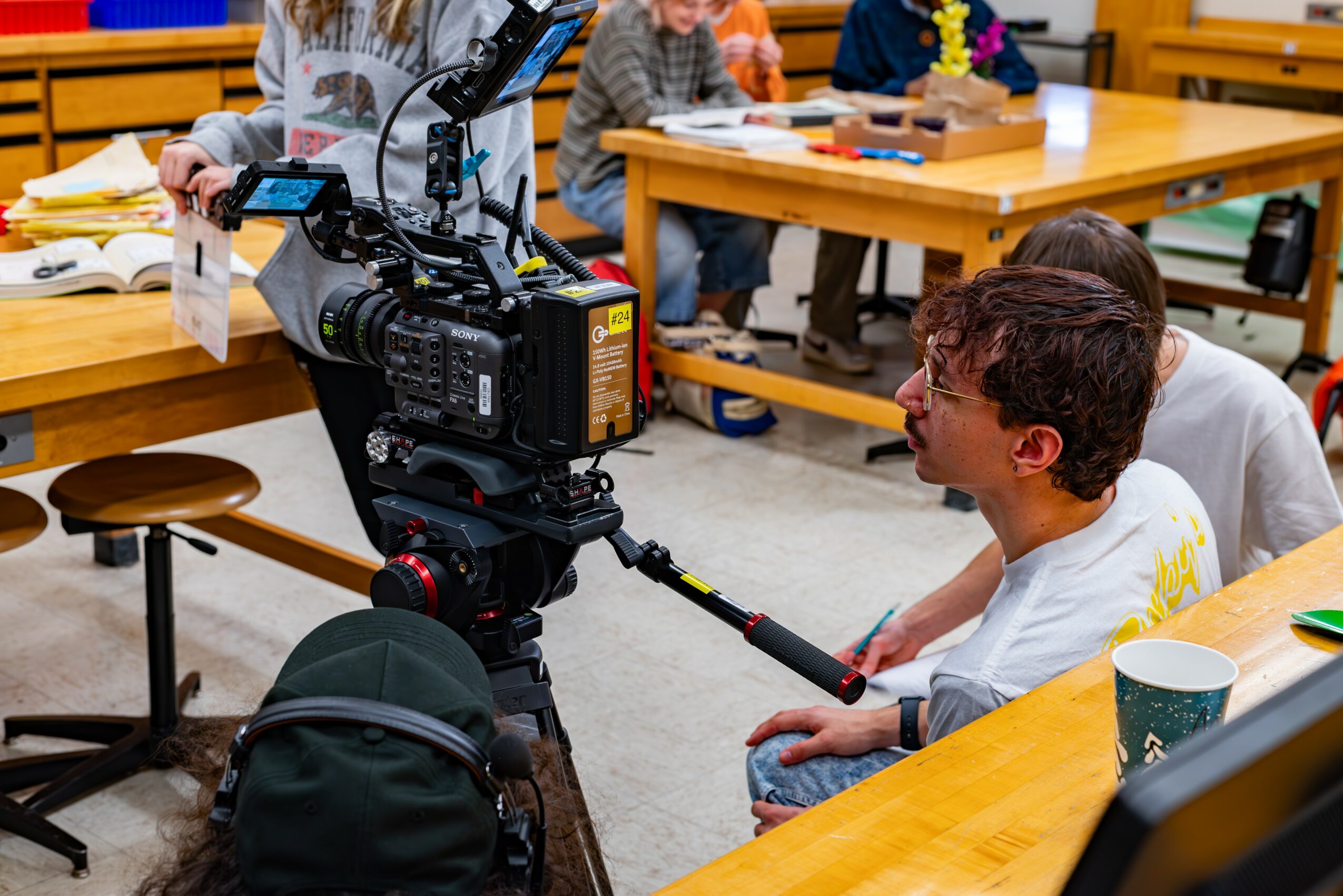 Person operating a professional video camera in a classroom with children at tables, some reading or writing, colorful decorations, and books.