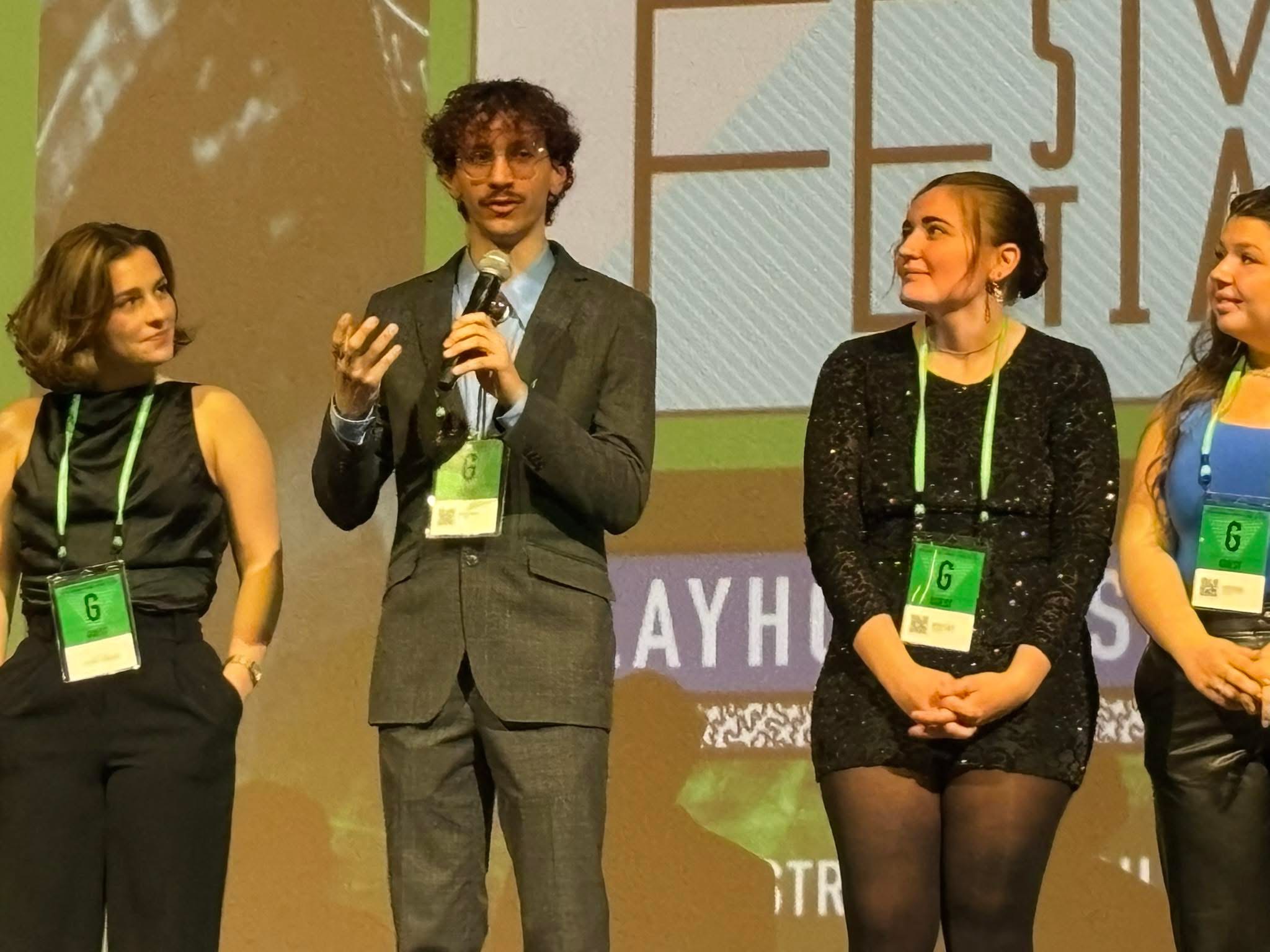 Four people on stage, one speaking into a microphone, three women listening, background with text and graphics.