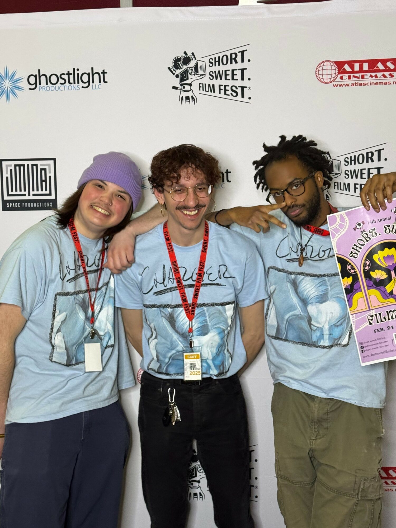 Three people standing together, smiling, with a white background and event banners behind them.