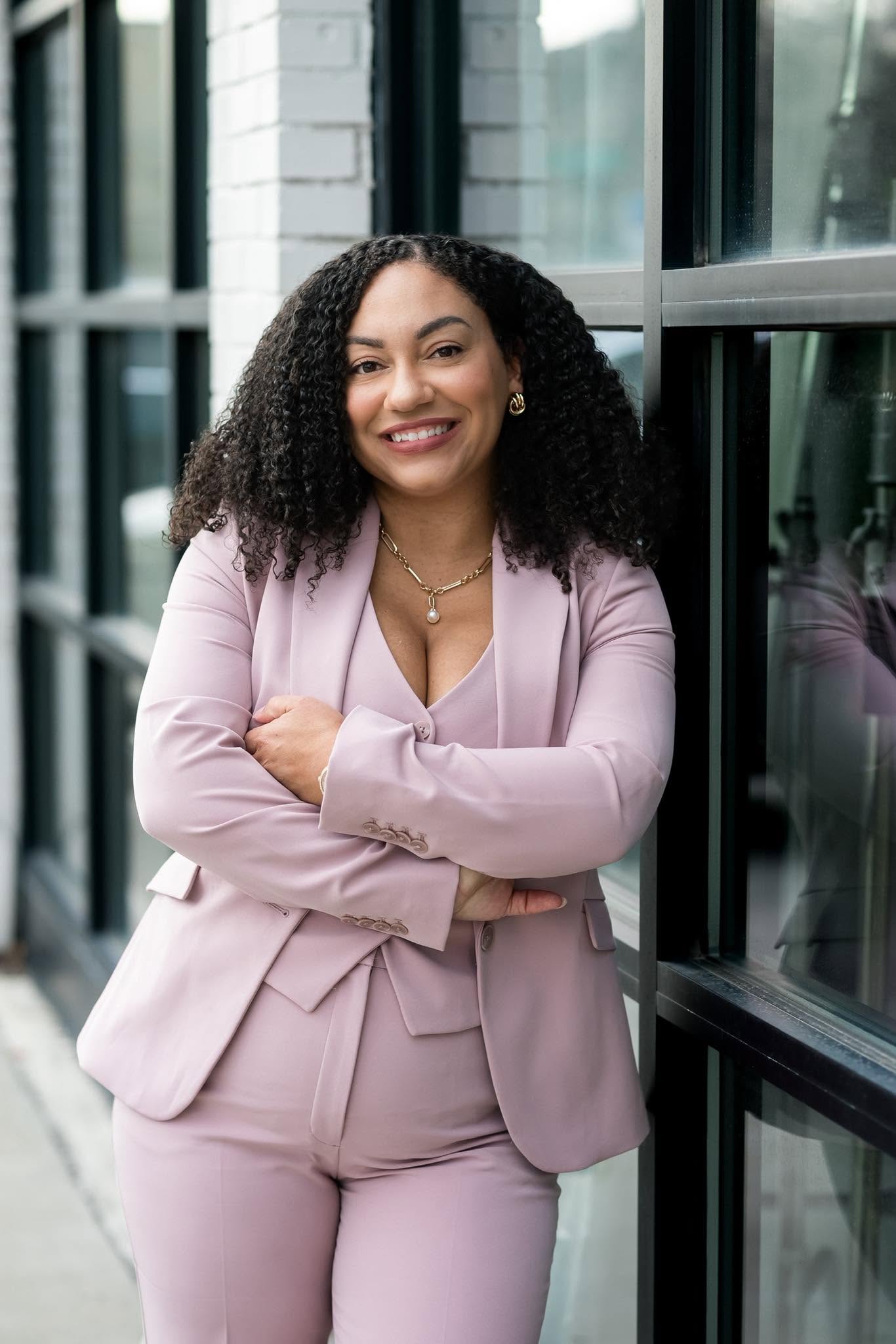 Smiling woman with curly hair in a pink suit standing outdoors near glass windows.