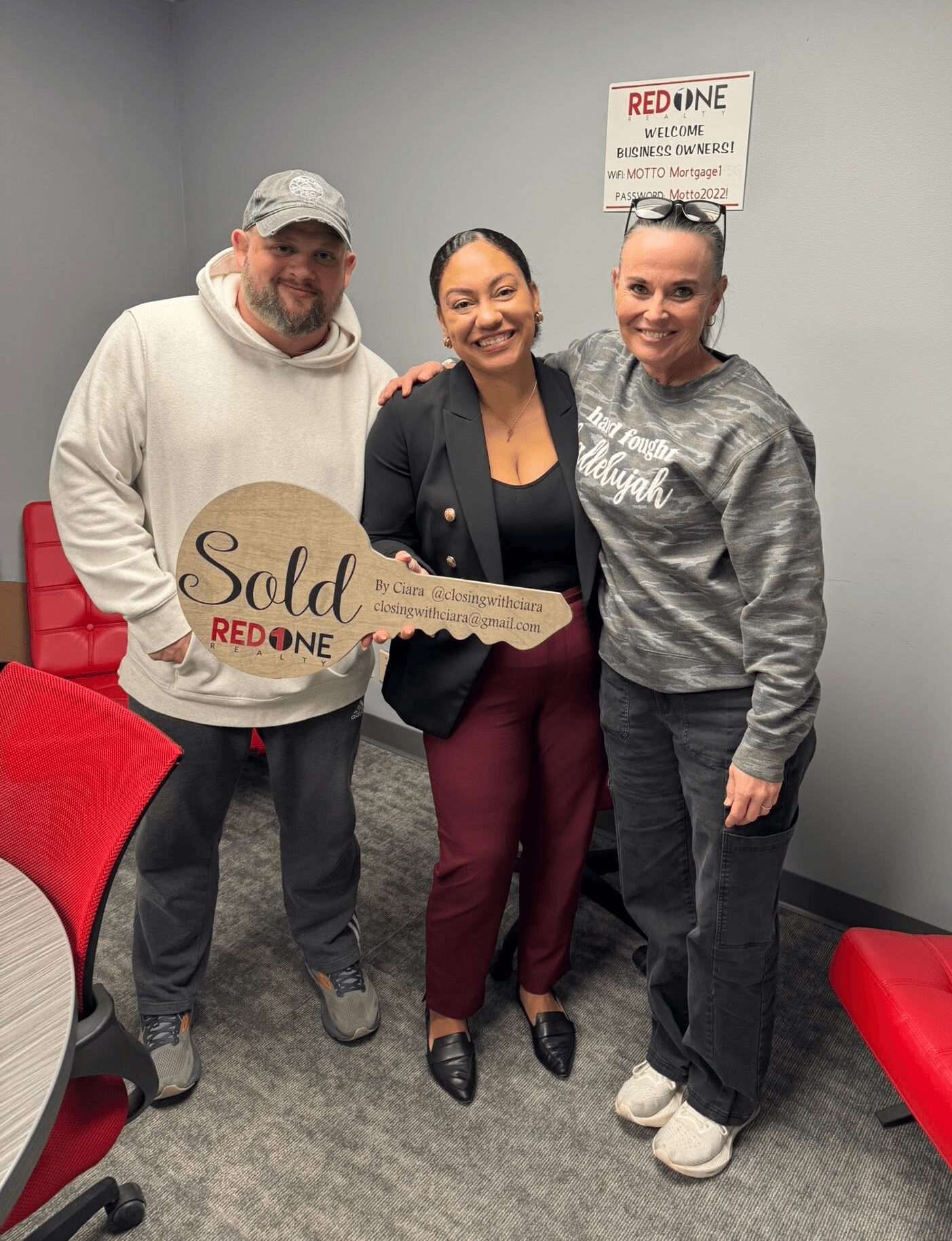 Three people standing indoors, smiling, with a sign that says 'Sold' and 'Red One' behind them.