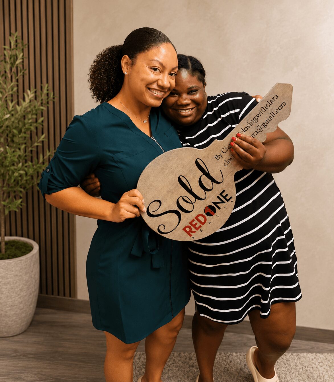 Two women smiling, one holding a large key-shaped sign with 'Sold' and 'REDDONE' written on it, standing indoors.