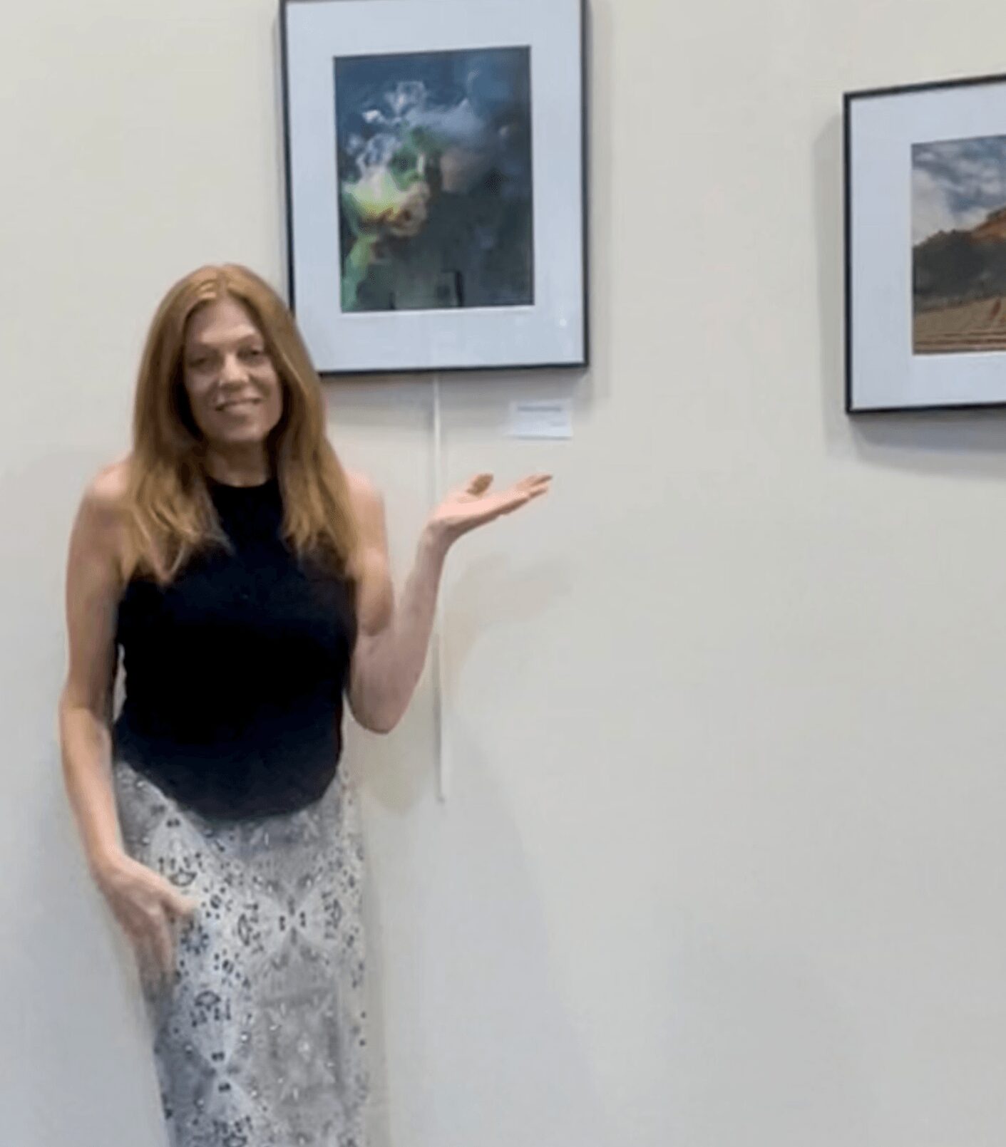 Woman with long hair in a black top and patterned skirt standing next to framed artwork on a white wall.
