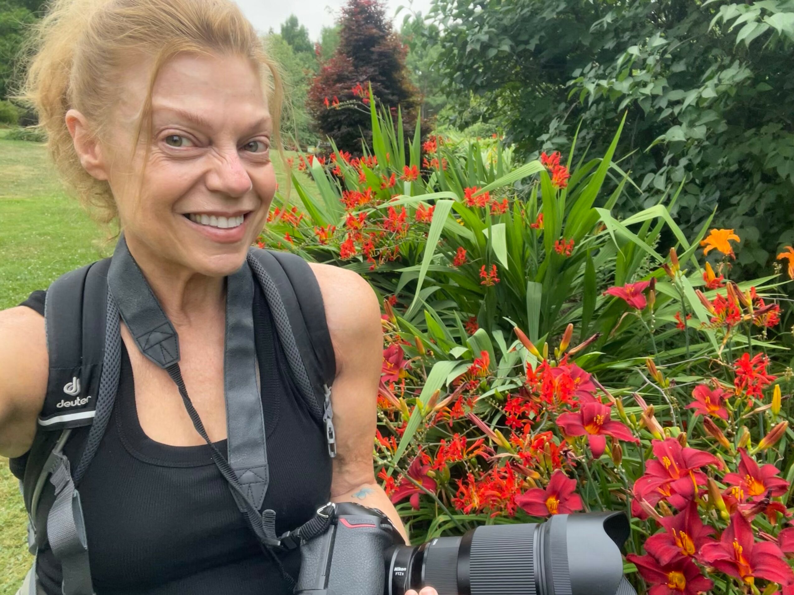 Smiling woman with a backpack holding a camera outdoors with colorful flowers and greenery in the background.