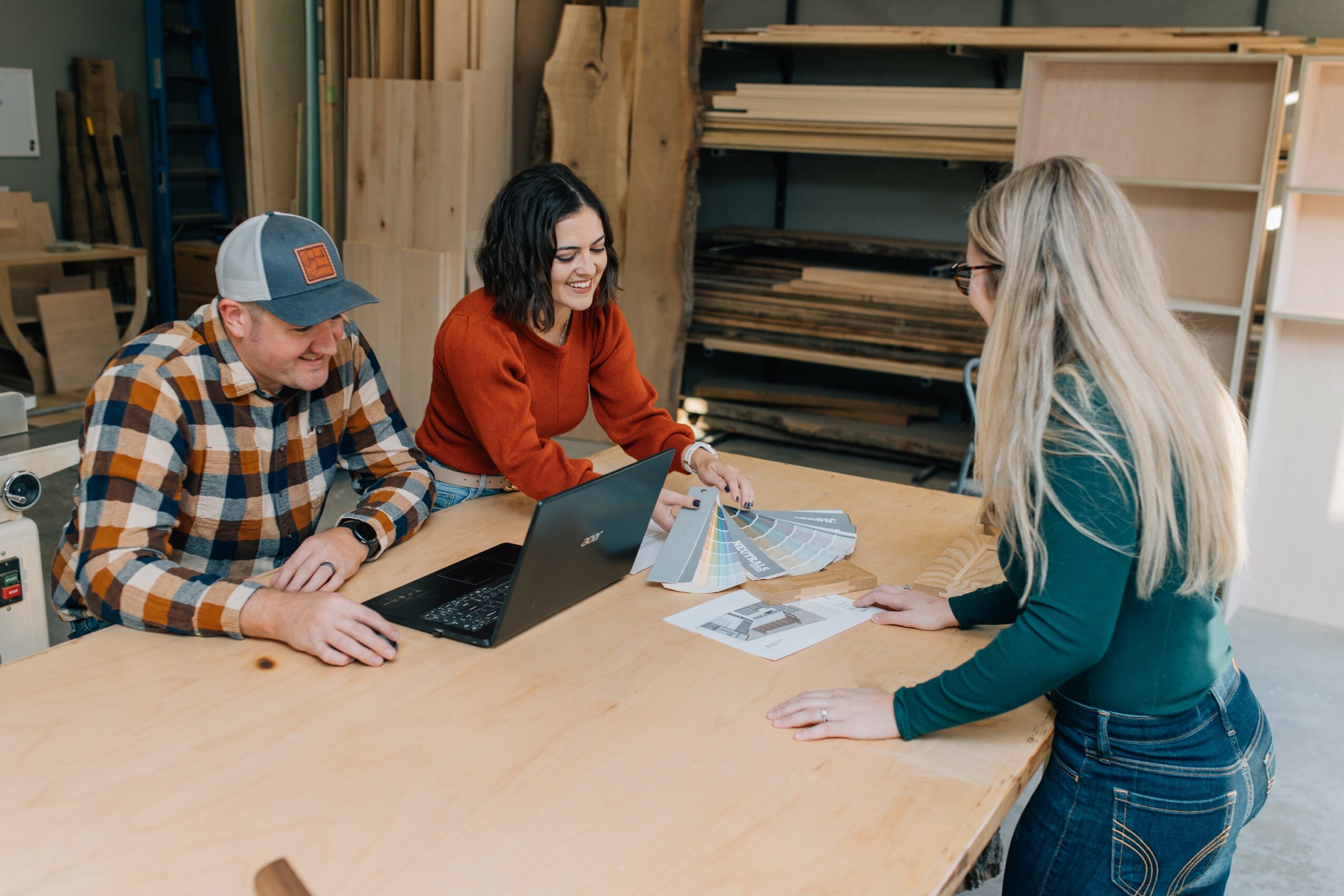 Three people sitting at a wooden table in a workshop, discussing documents and using a laptop, with shelves and wood in the background.