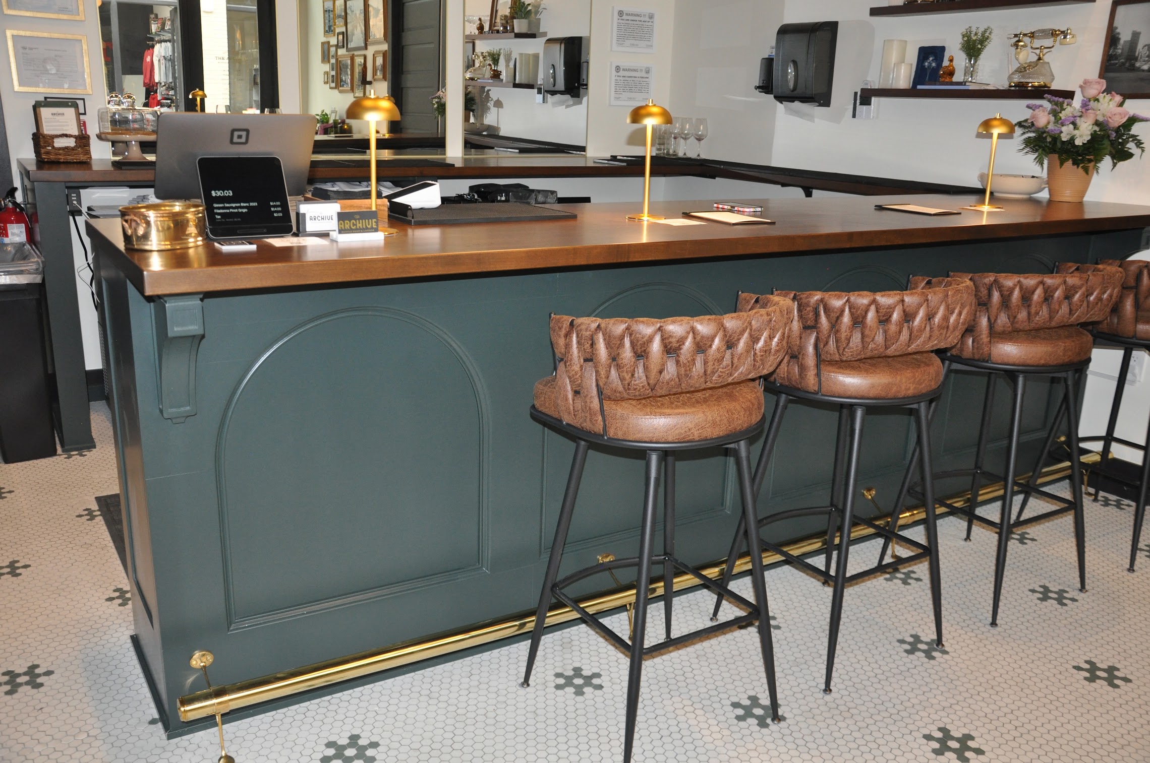 Bar counter with teal base, brown leather stools, and a wooden top in a cafe setting, with lamps and decor in background.