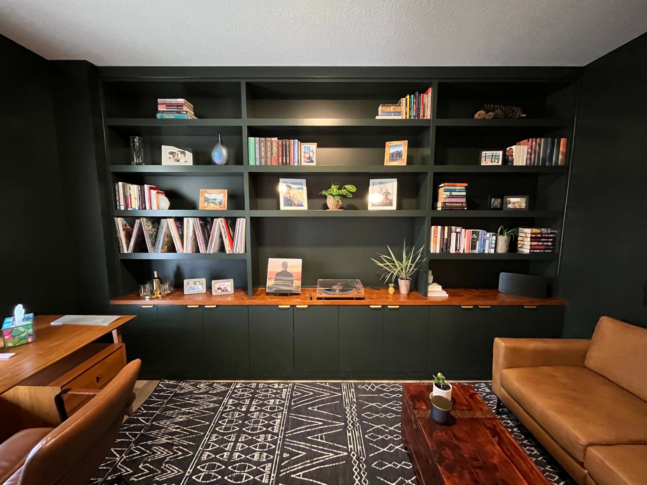 Living room with black bookshelf, brown sofa, and wooden furniture, patterned rug, and decorative items on shelves.