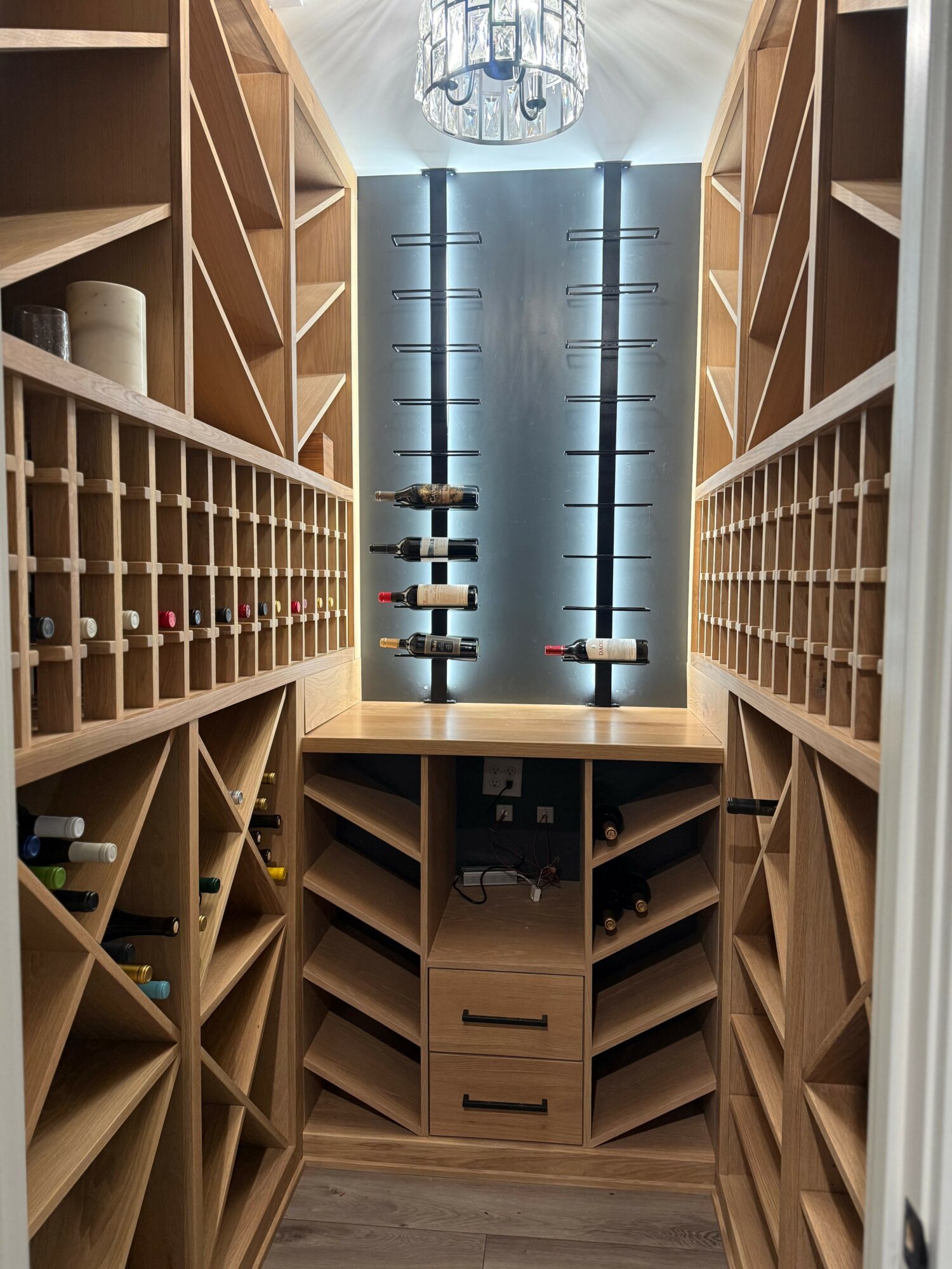 View of a wine cellar with wooden shelves, bottles, and a central table with wine bottles, illuminated by a ceiling light.