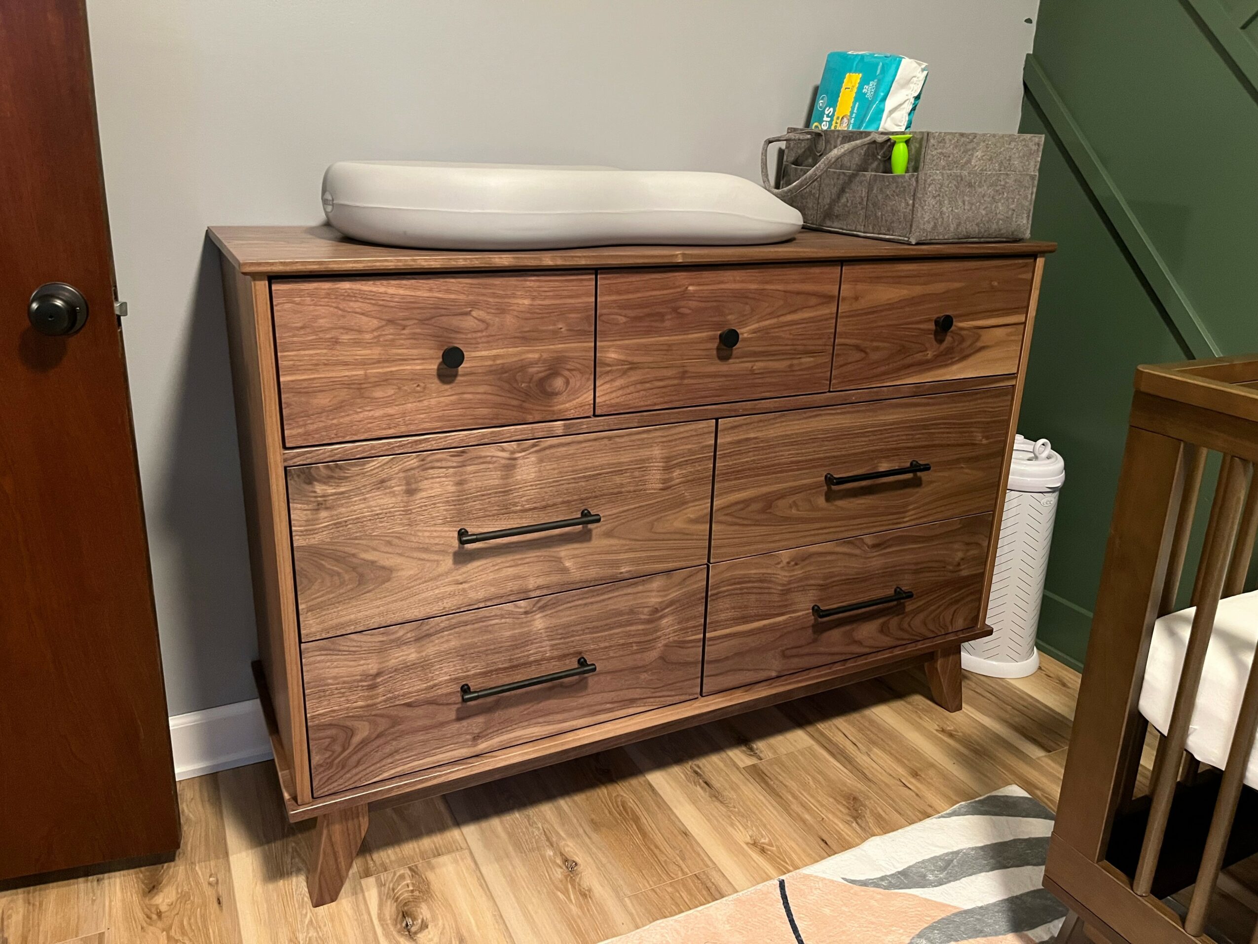 Wooden changing table with six drawers, a white changing pad on top, and a gray storage box with baby supplies, in a nursery.