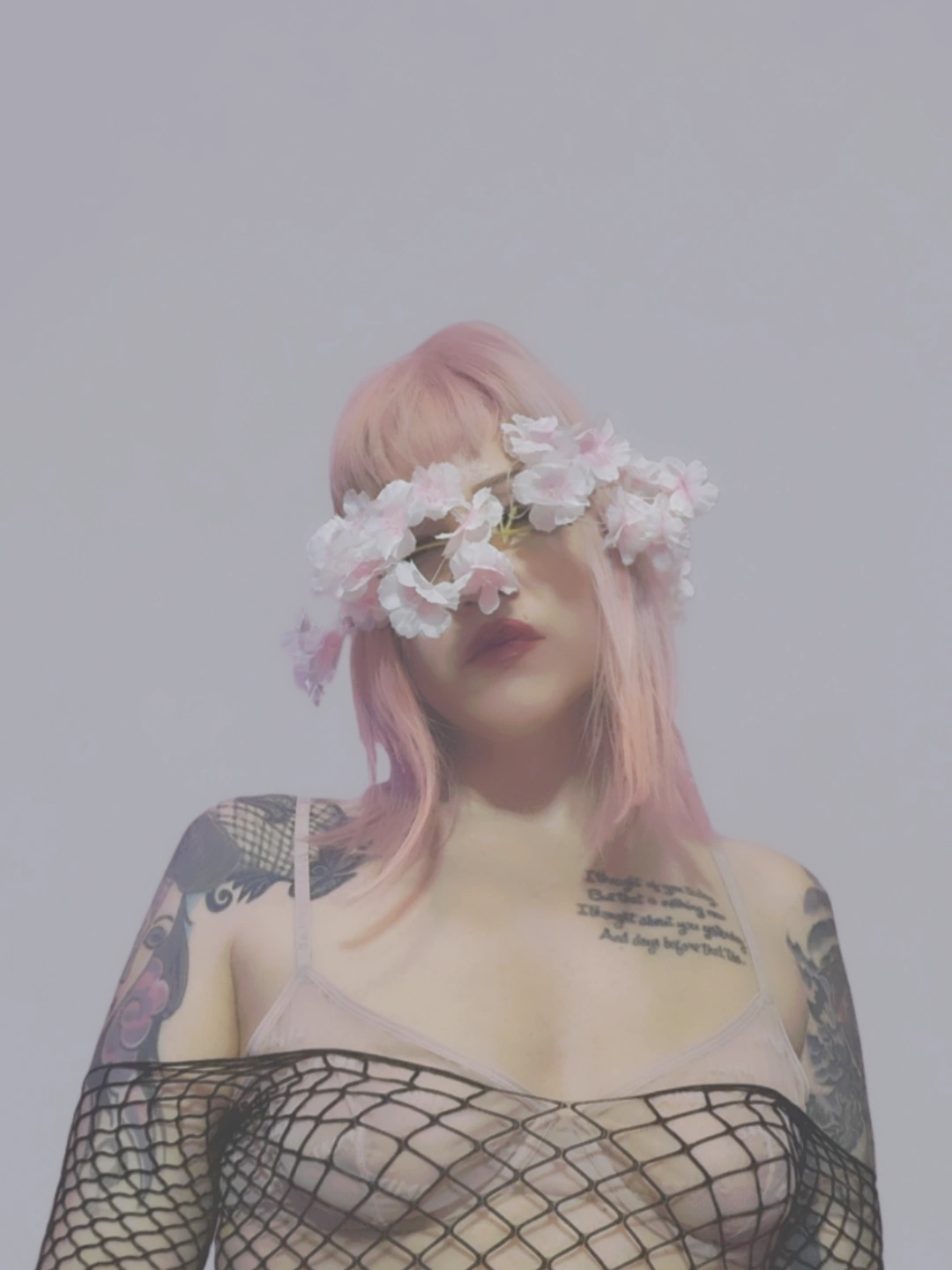 Person with pink hair wearing a flower crown and a black netted top, with tattoos on shoulders and chest.