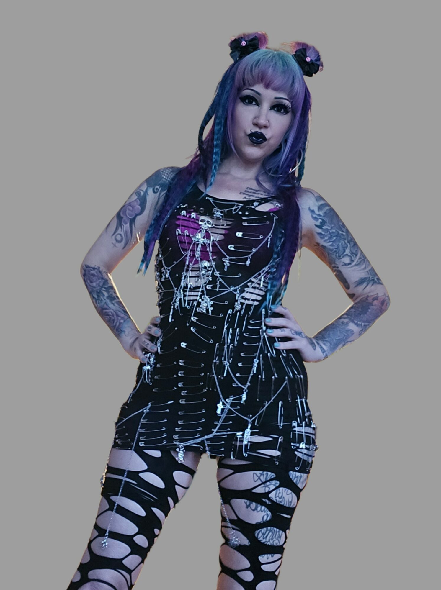 Person with purple hair, tattoos, wearing a black dress with white patterns and ripped leggings, standing with hands on hips.
