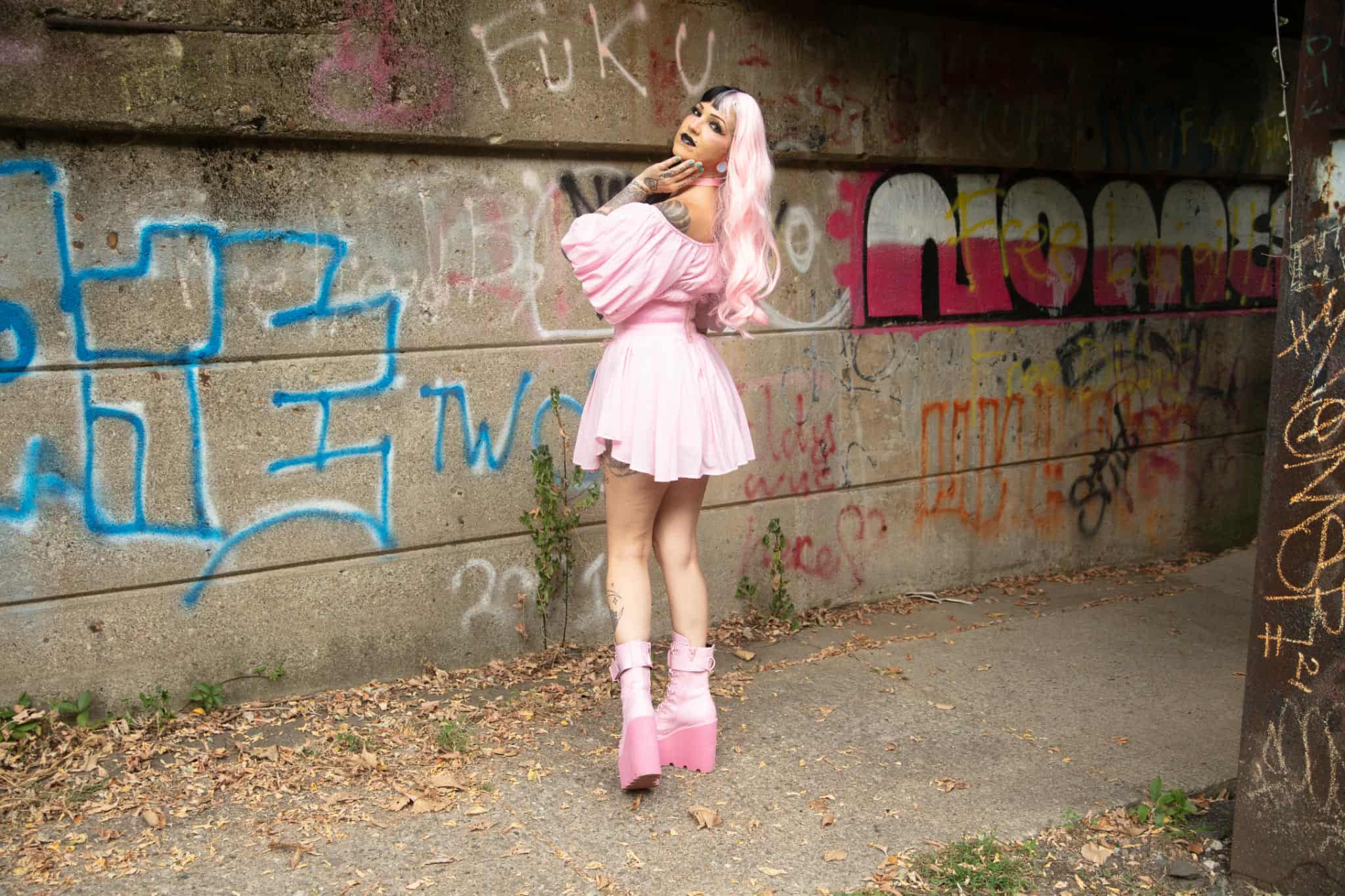Person in pink dress and platform shoes standing against graffiti-covered wall, with one hand on head, outdoors.